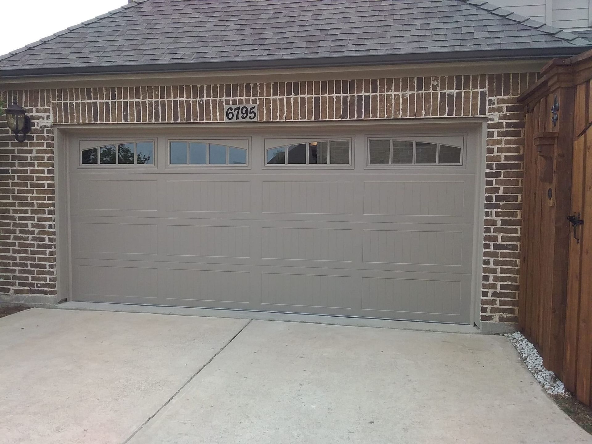 A tan garage door with a row of small windows at the top, installed in a brick house with a dark shingled roof.