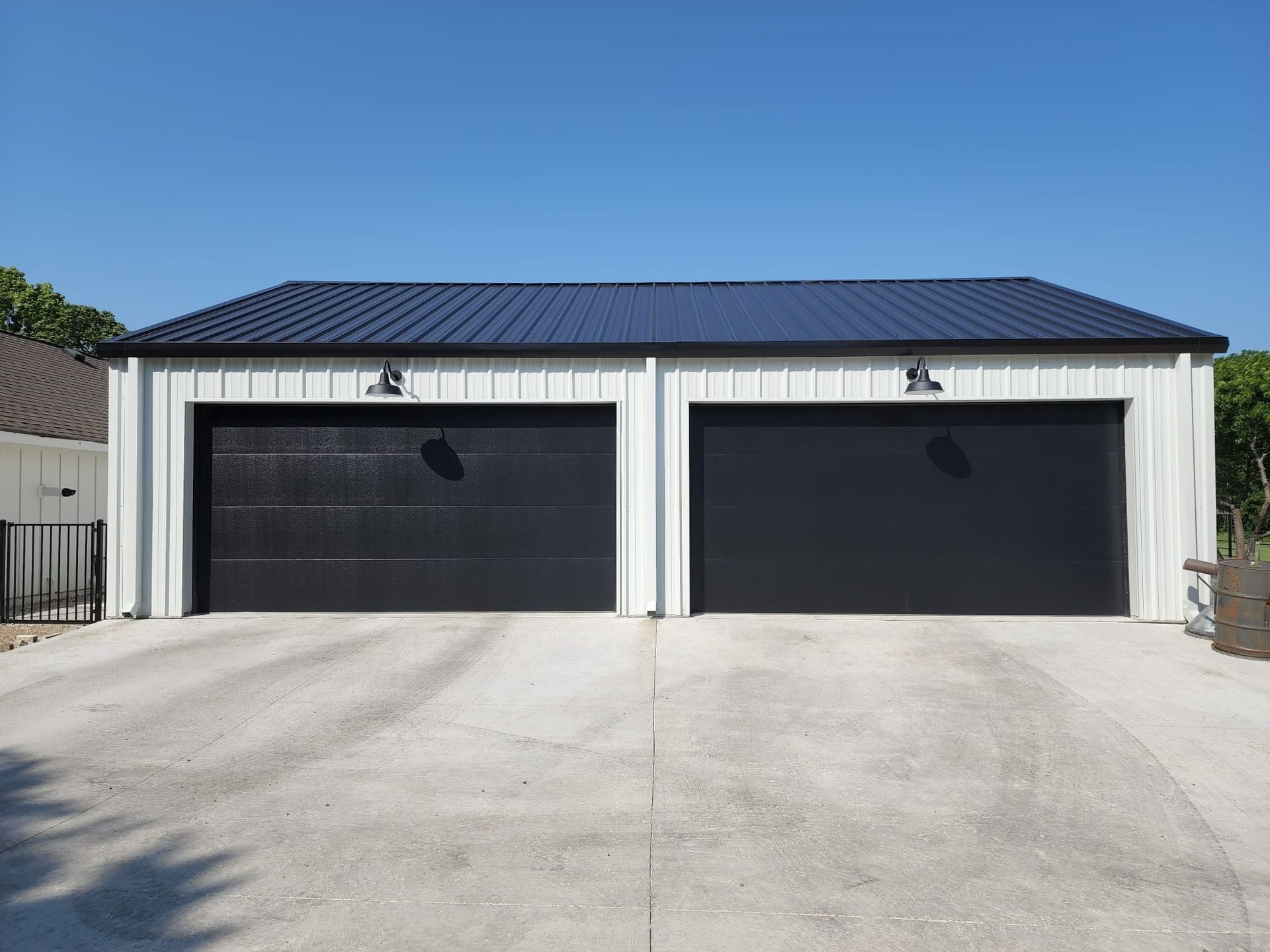 A detached, two-car garage with vertical white siding, a dark blue metal roof, and two solid black garage doors.