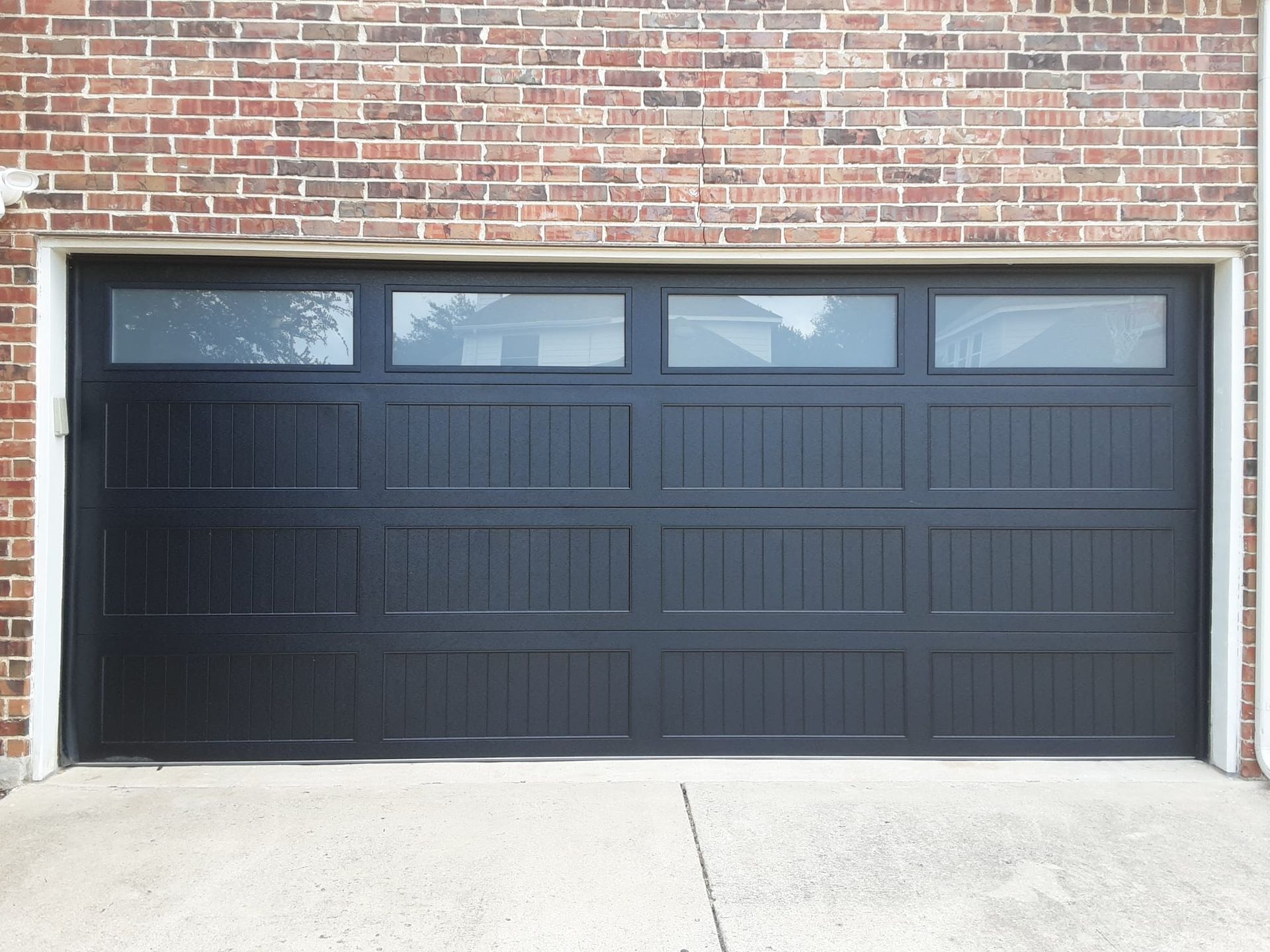 A black, four-section garage door with top-row windows, set against a brown brick exterior.