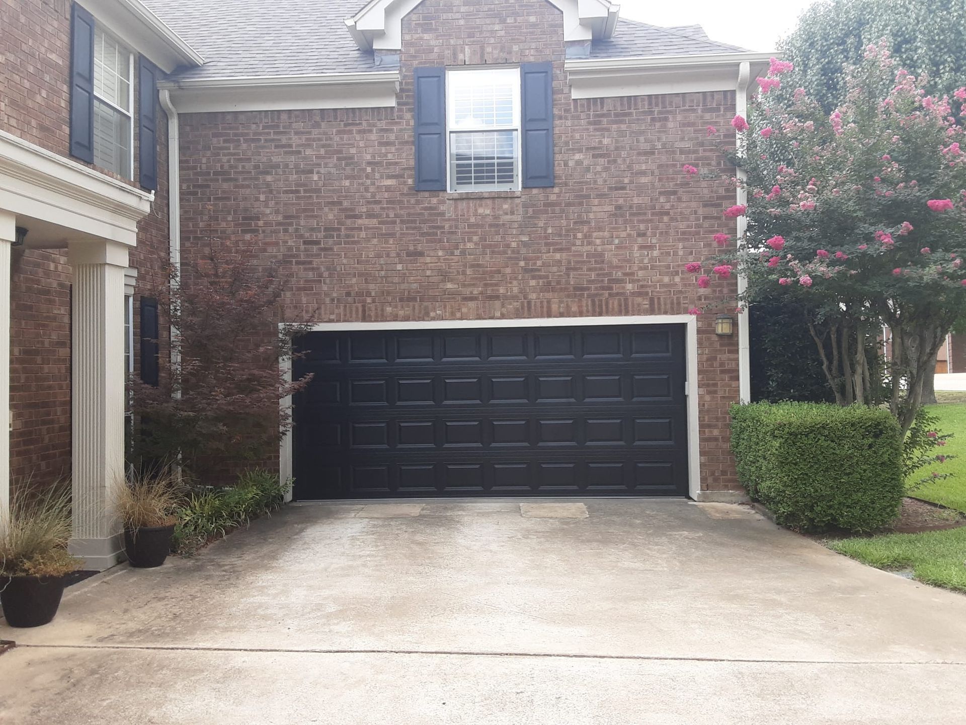 A brick home with a black garage door and a green tree and shrubs in the front yard on a concrete driveway.