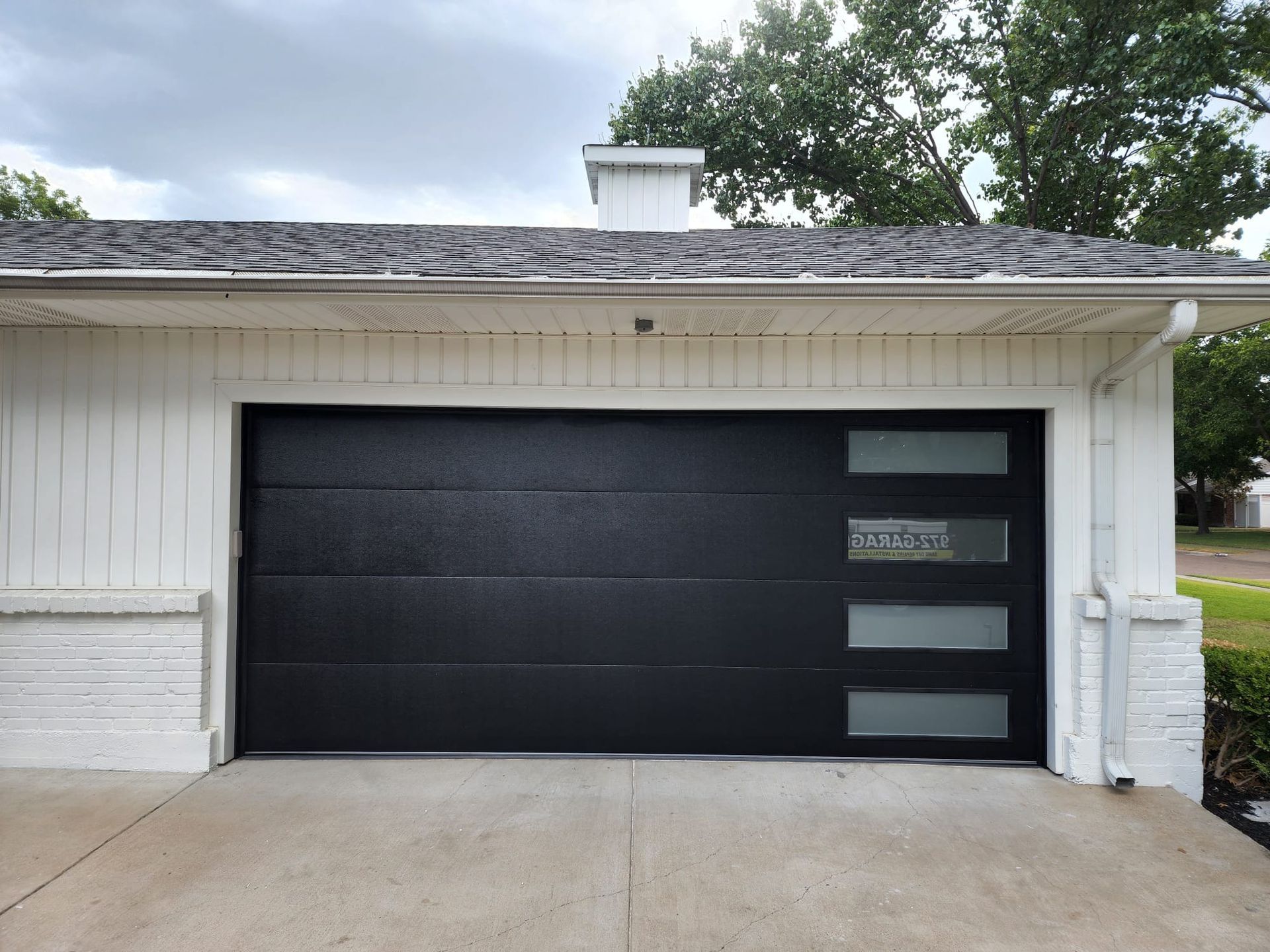 A black, modern-style garage door with a vertical row of four frosted glass windows on the right, set in a white garage.