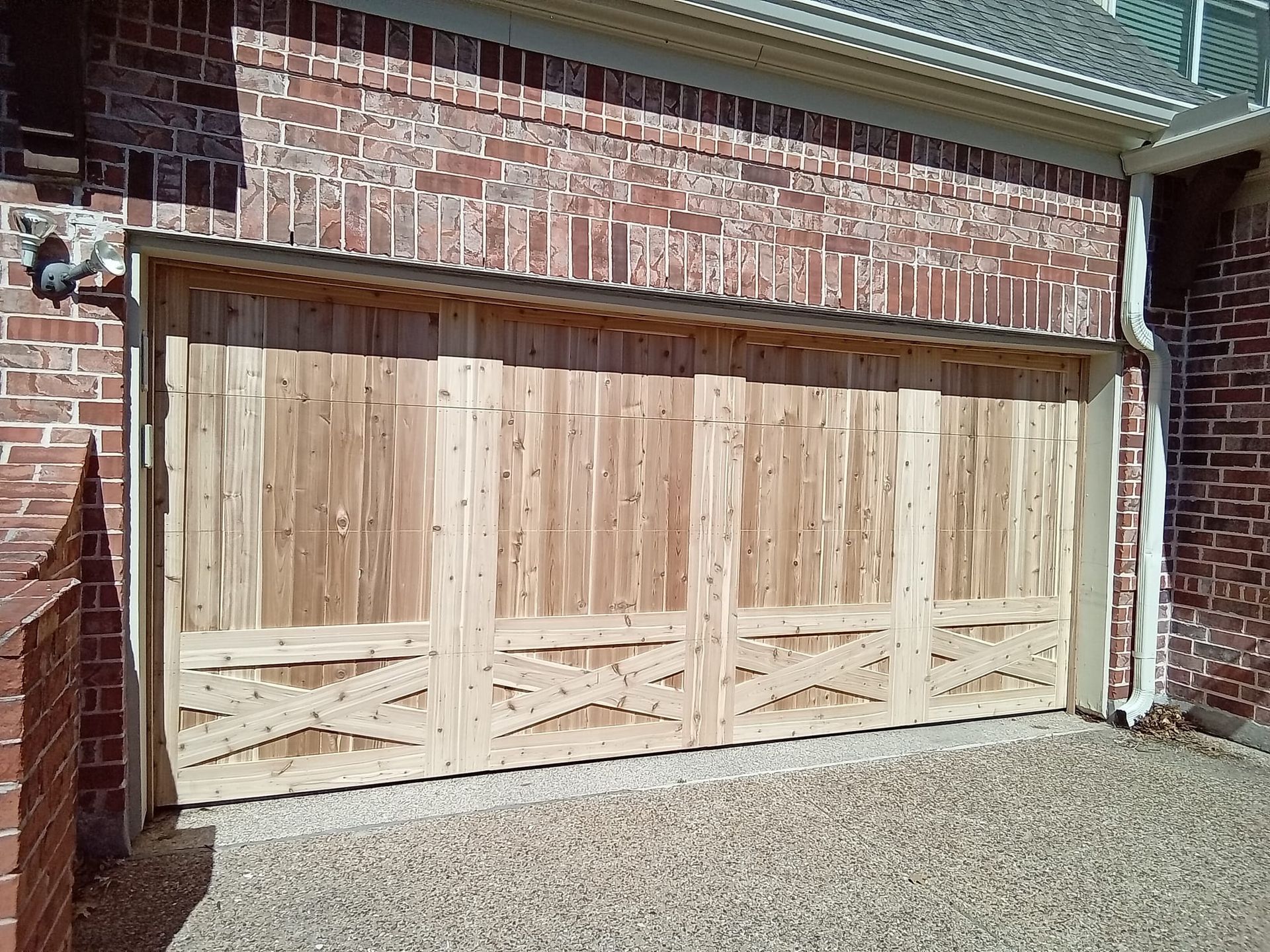 A light-toned wood garage door with a decorative X-brace design set into a reddish-brown brick house exterior.