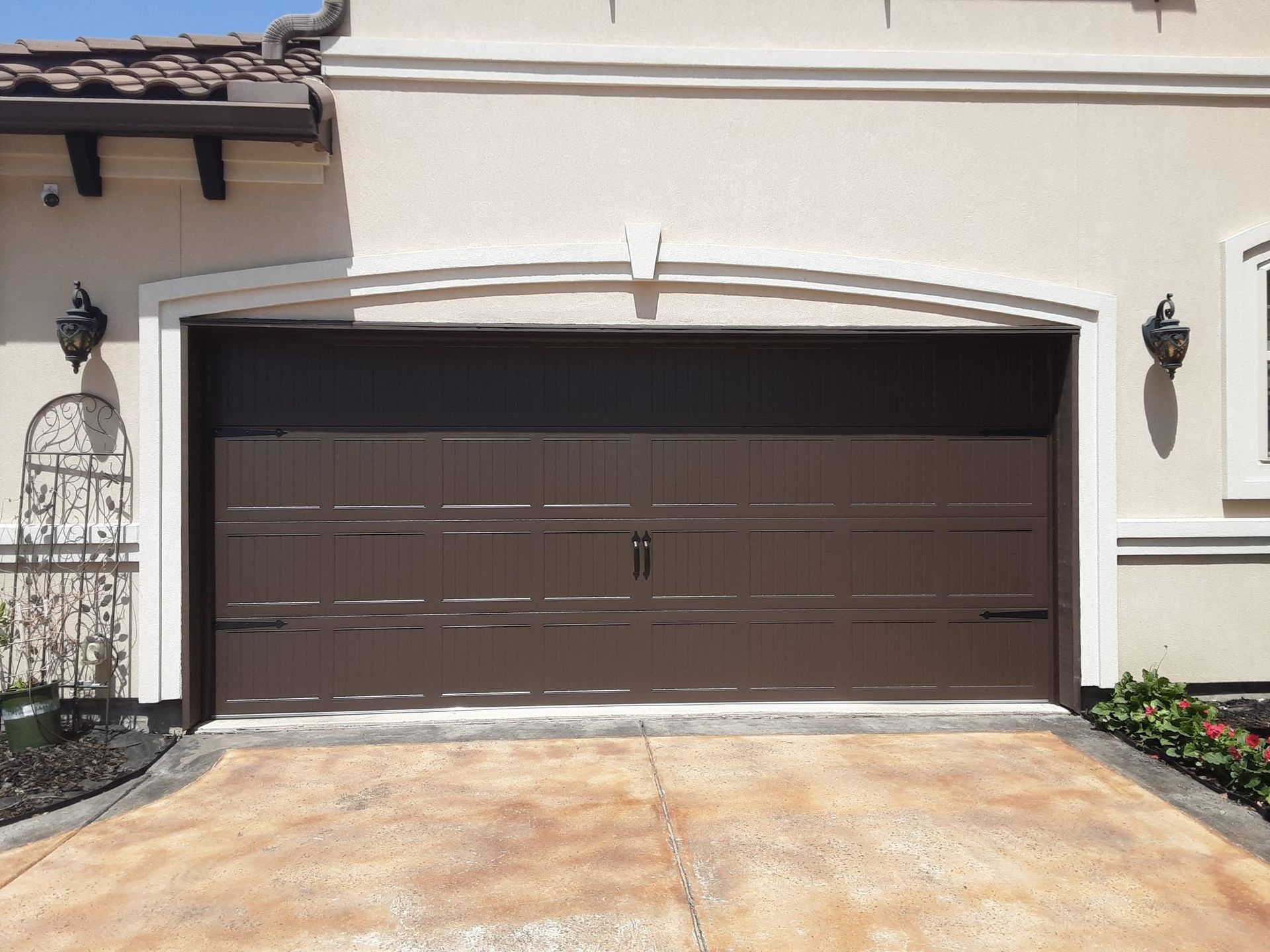 A brown, paneled double garage door set in a beige home exterior with arched trim, black light fixtures, and a driveway.