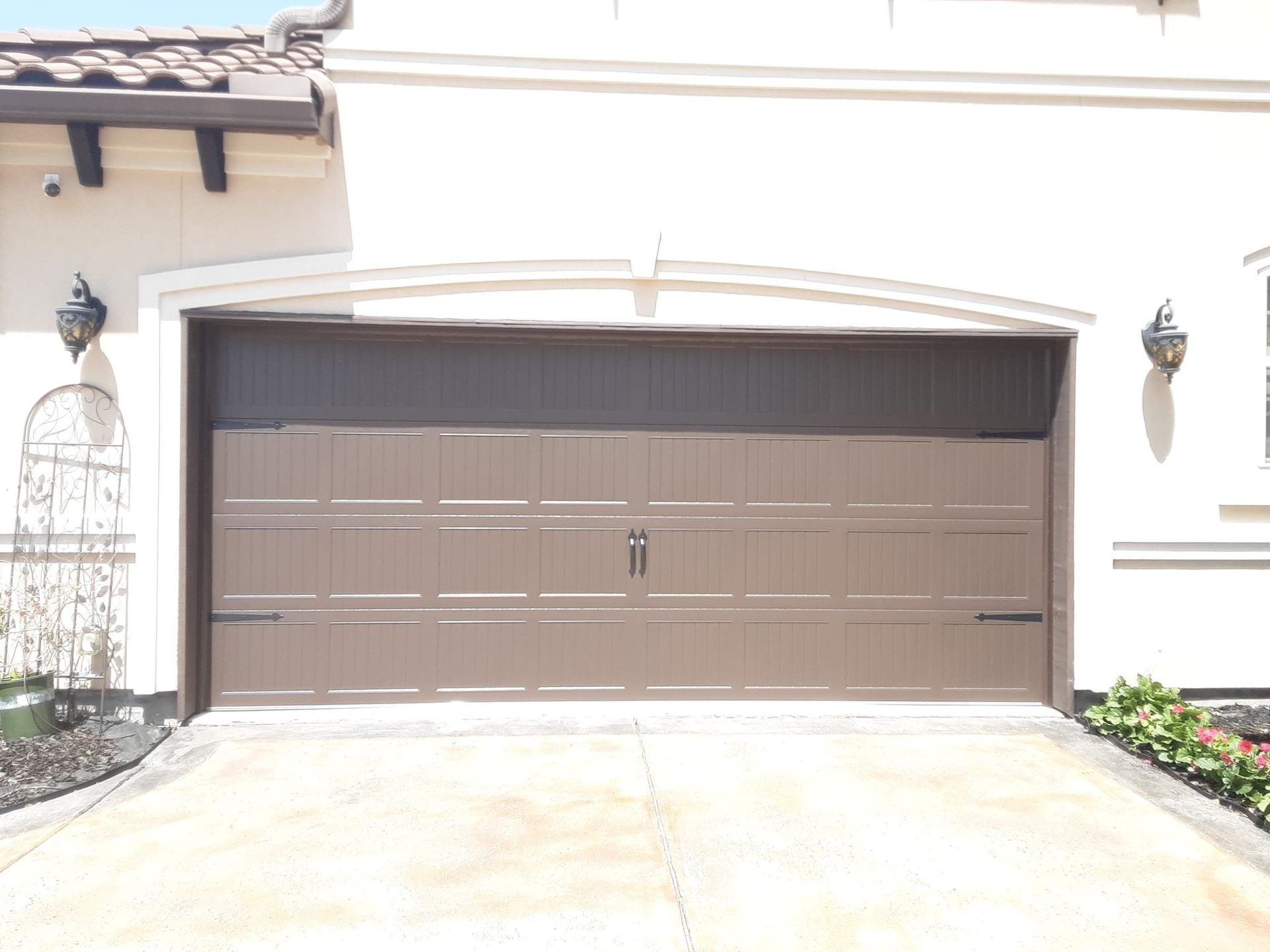 A dark brown, rectangular garage door with horizontal panels, centered handles, and side sconces on a light-colored home.