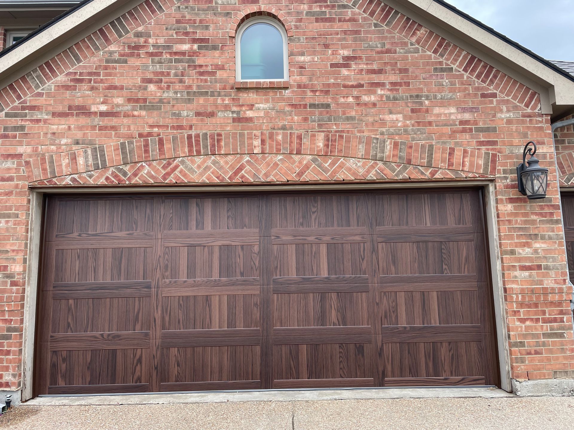 Dark brown, paneled garage door set in a red brick wall with an arched brick header and small arched window above.