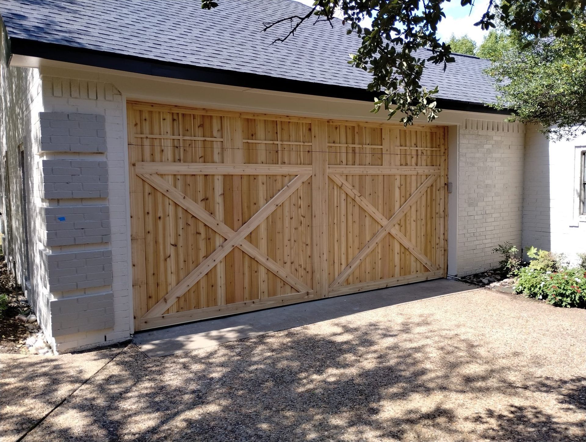 A light-colored wooden carriage-style garage door with an X-brace design set in a white brick wall.