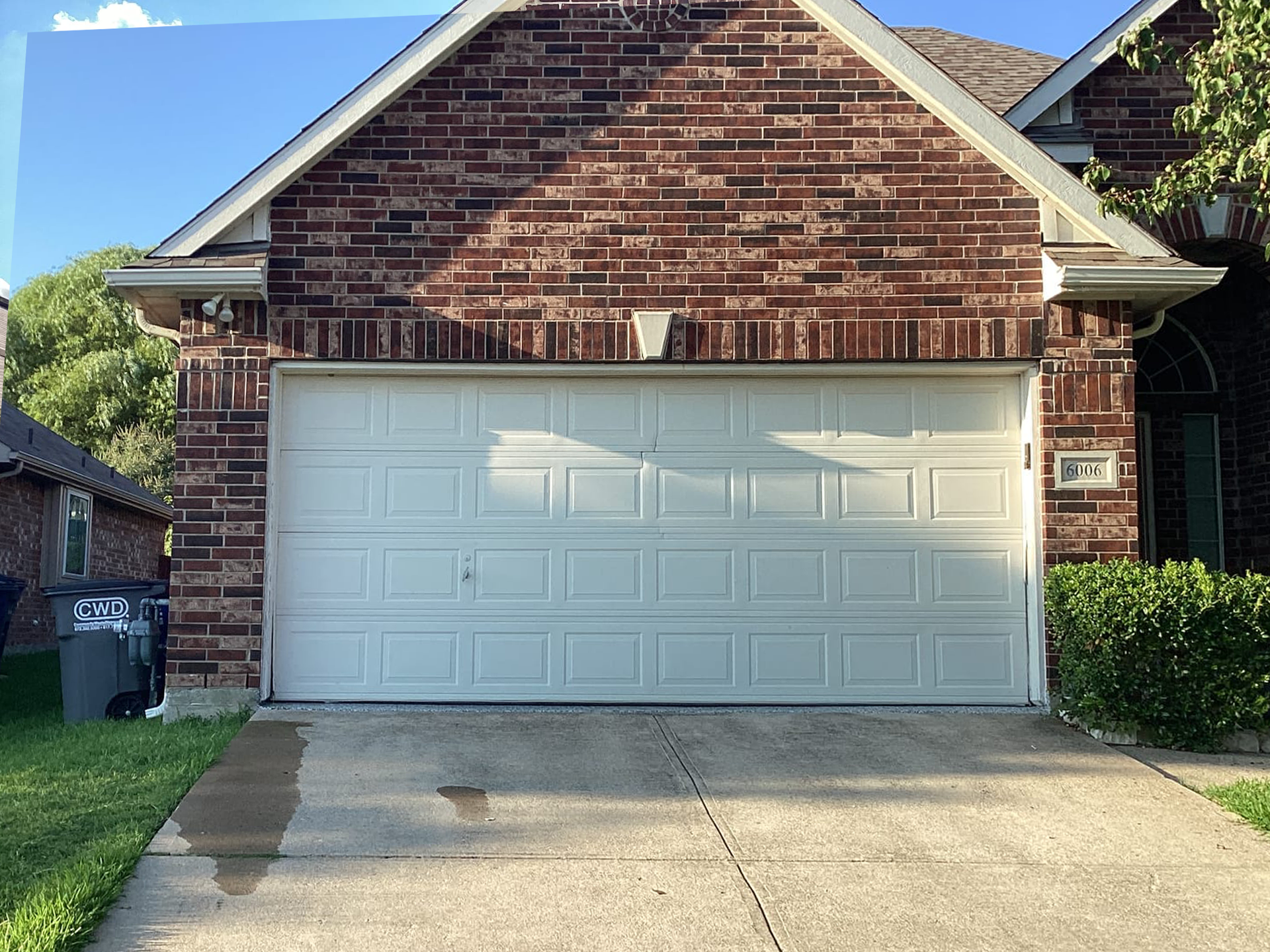 A brick house with a white garage door, trees, and trash cans.