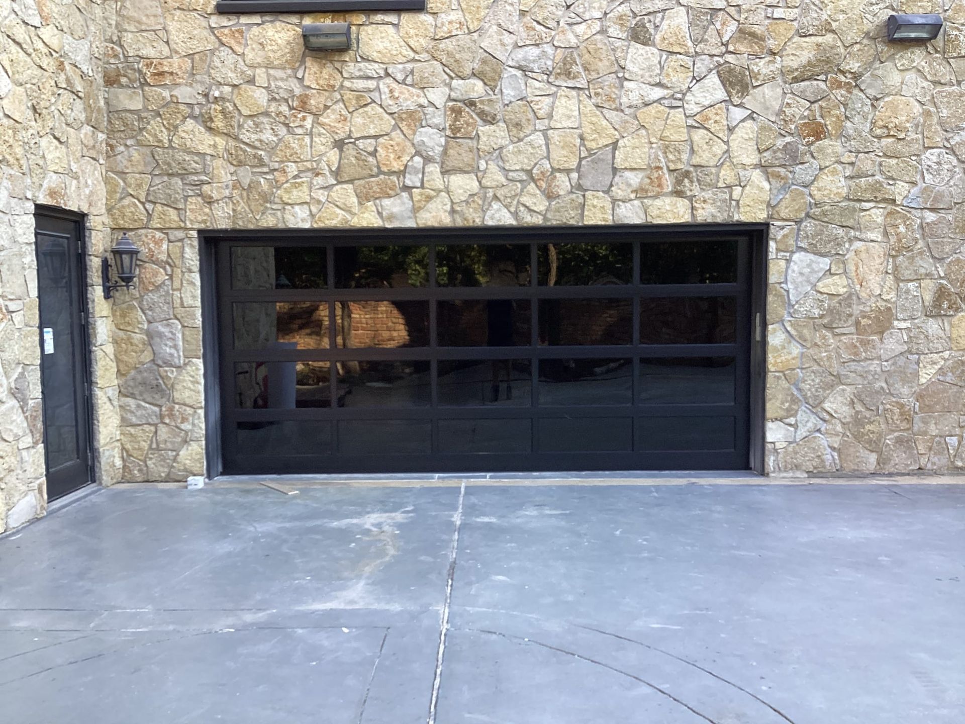 A modern, black-framed glass garage door set into a textured, light-colored stone wall above a gray concrete driveway.