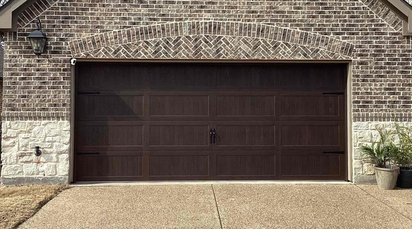 Brown brick home with a closed garage door. There is a gravel driveway and plants on the right.
