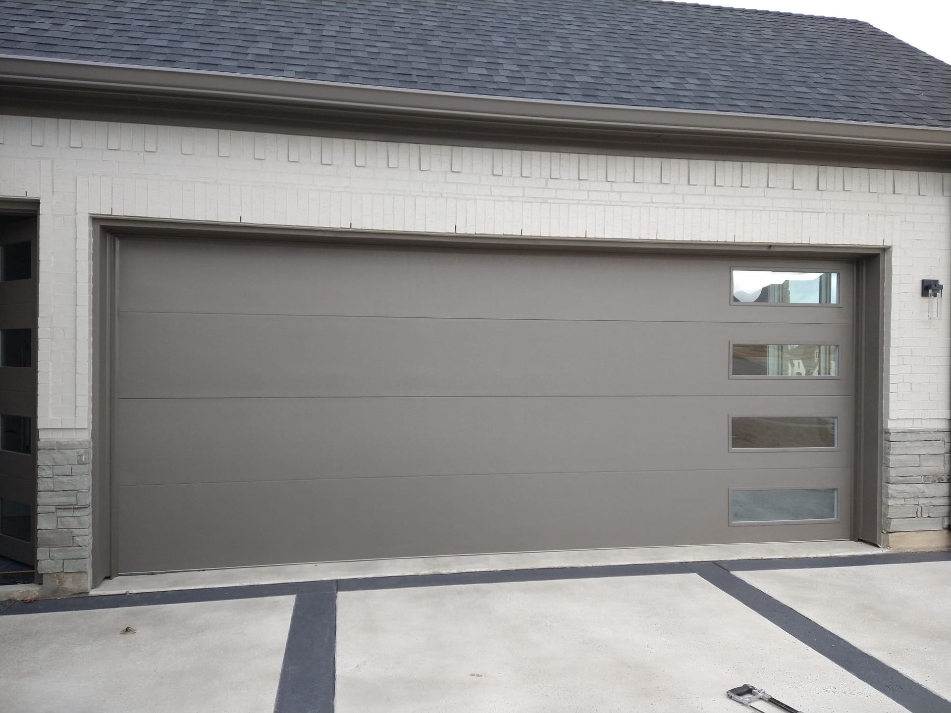 A taupe garage door with four vertical stacked rectangular windows on the right side, set in a light-colored stone home.