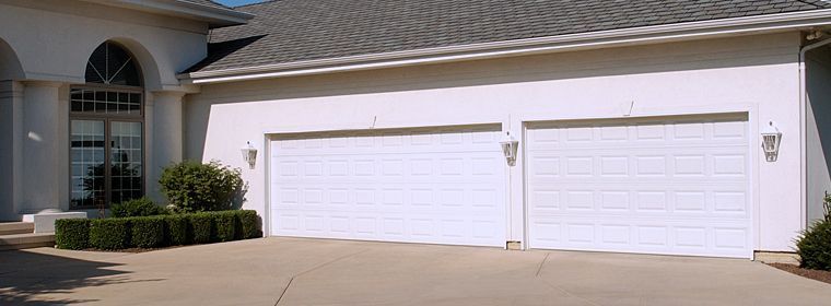 A residential house exterior featuring a beige stucco facade, a dark gray shingled roof, and two white garage doors.