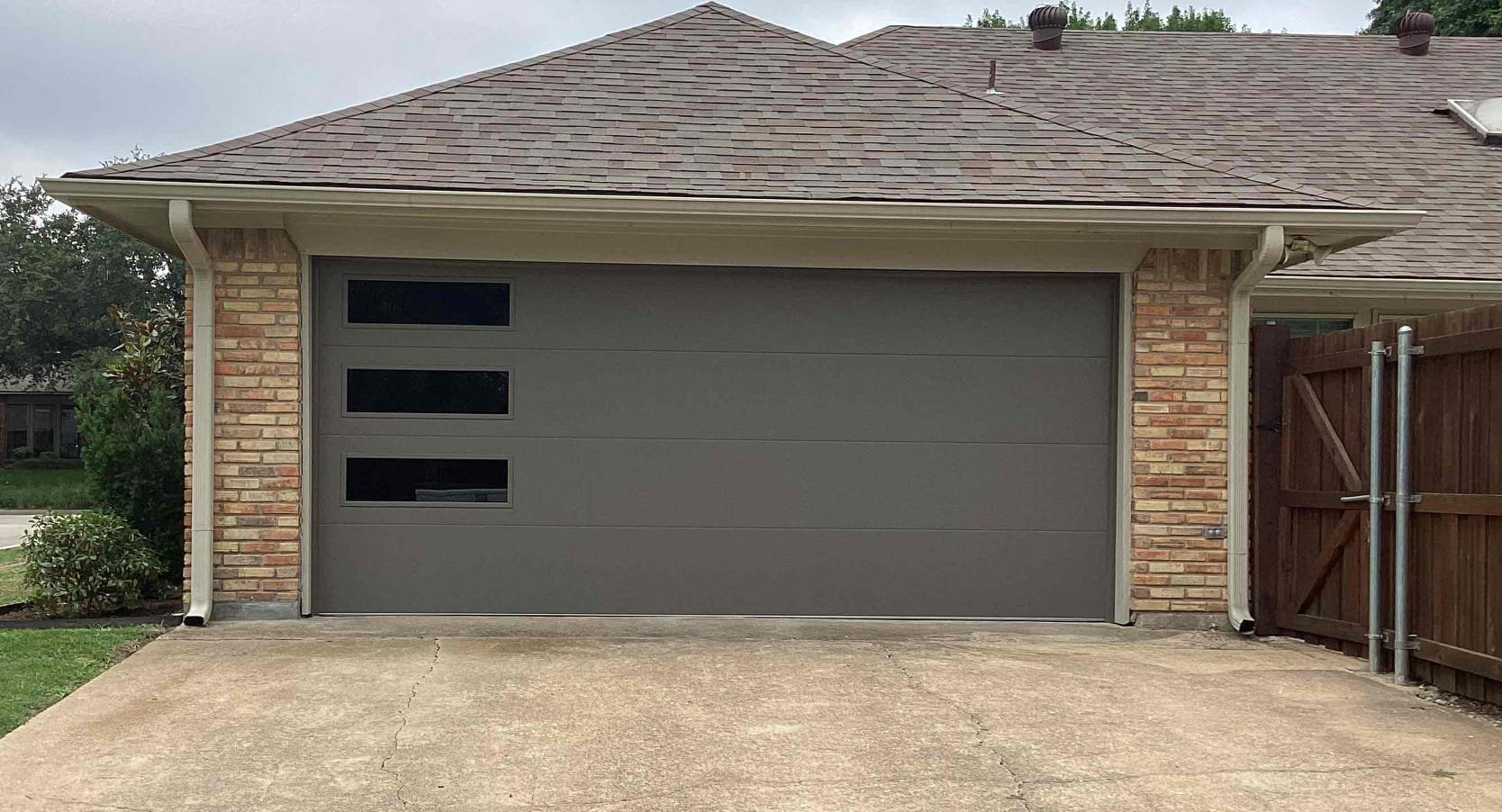 Gray garage door with three horizontal windows, tan stone facade, and brown roof.