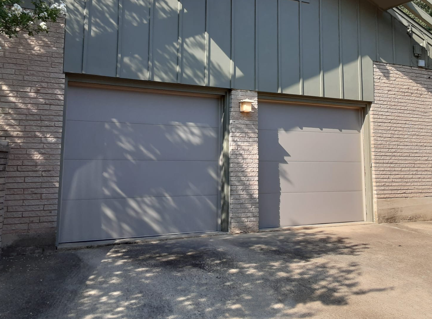 Two gray garage doors in a brick and green-paneled building with sunlight casting shadows on the driveway.