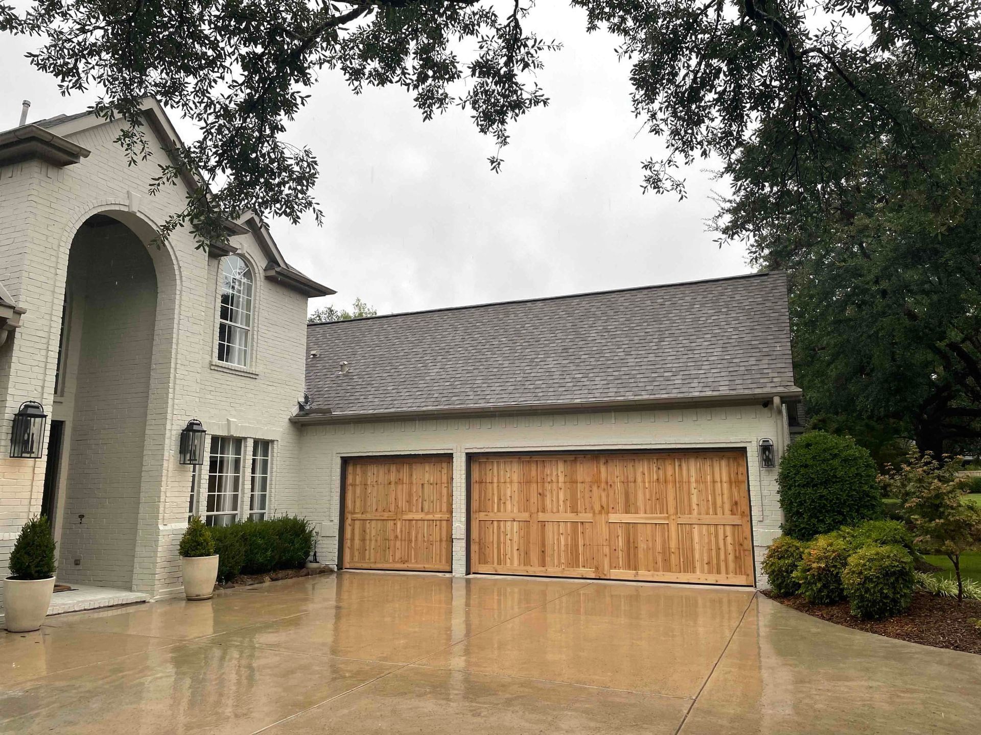 A two-story white brick house with an arched entry and a two-car garage with light wood doors under a cloudy sky.