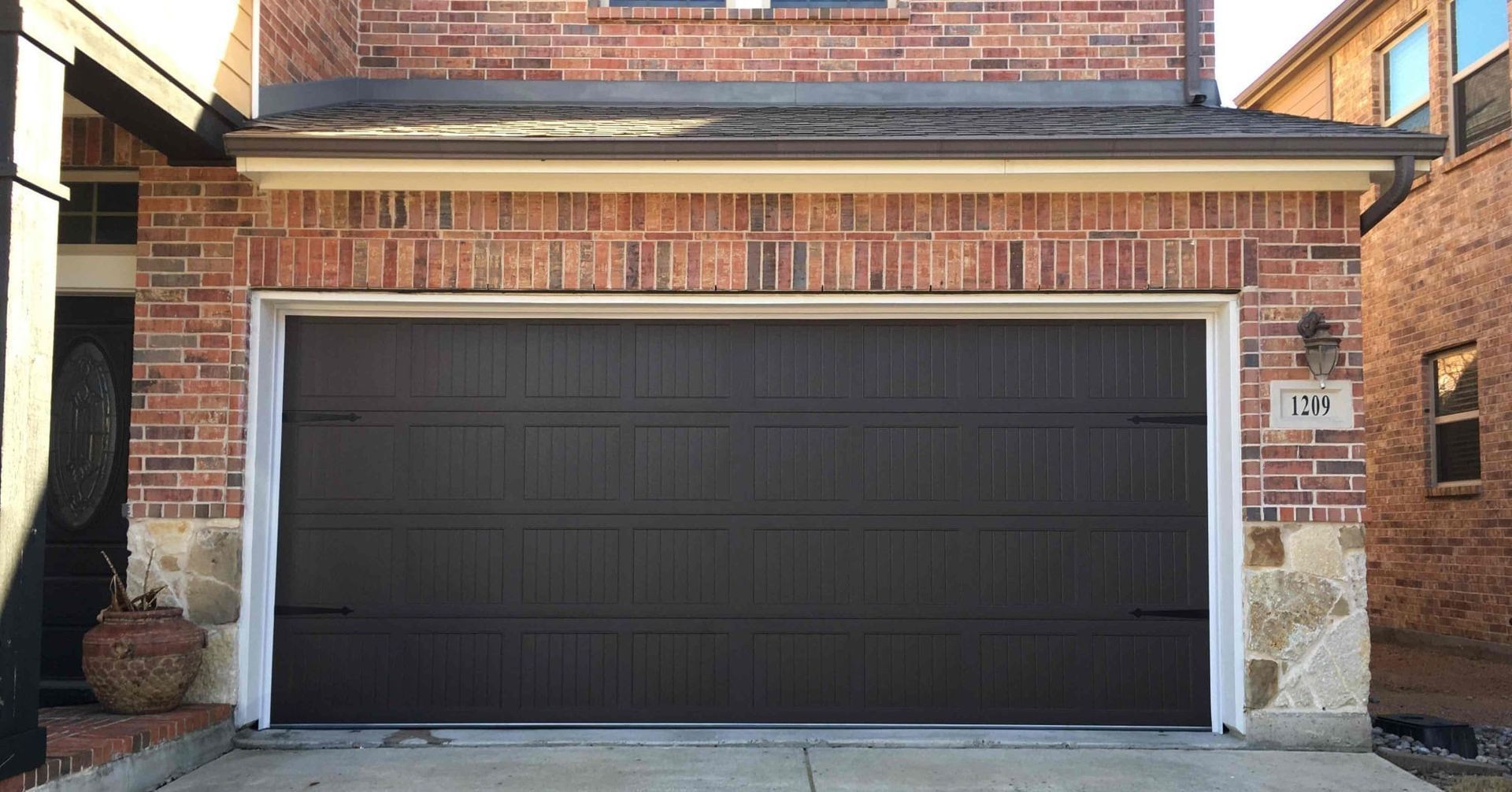 A dark brown, paneled garage door installed in a red brick suburban home with a white frame.