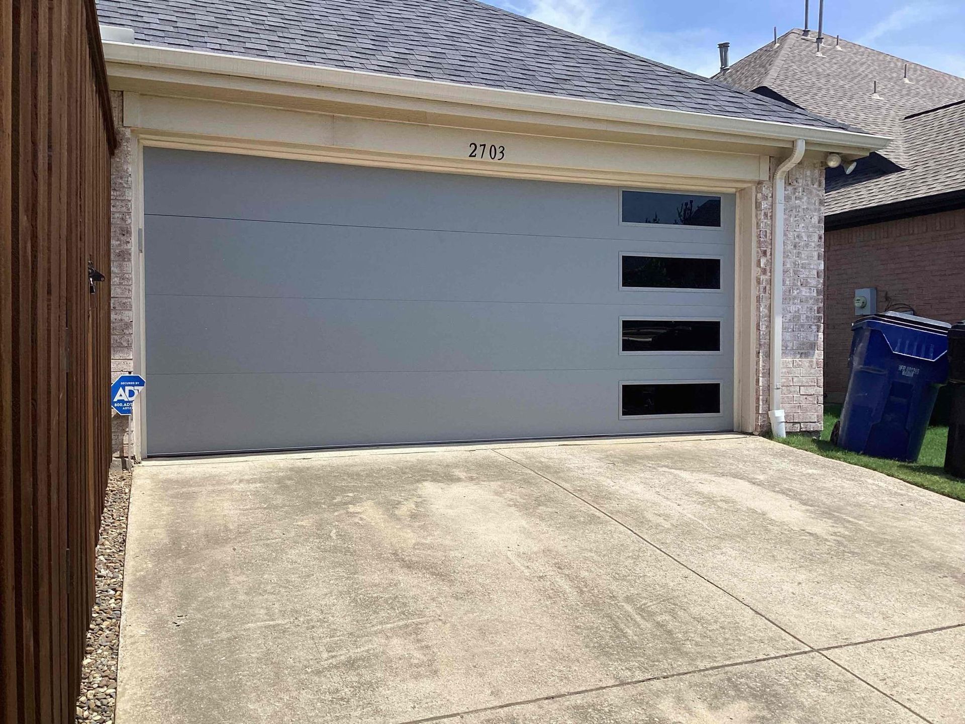 A gray garage door with a vertical row of four rectangular black windows on a residential home with a concrete driveway.