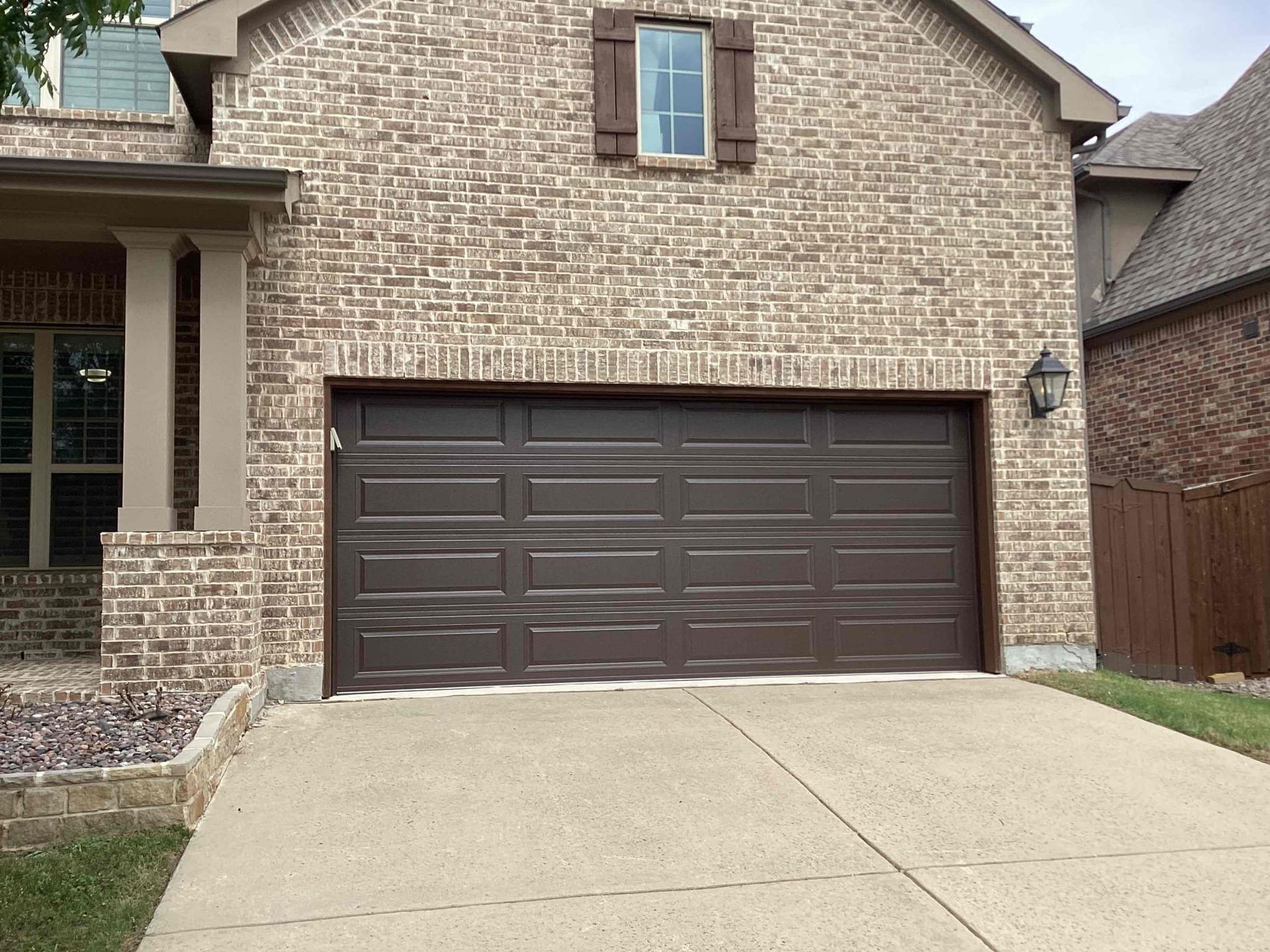 A brown garage door on a brick suburban home with a small window above and a concrete driveway.
