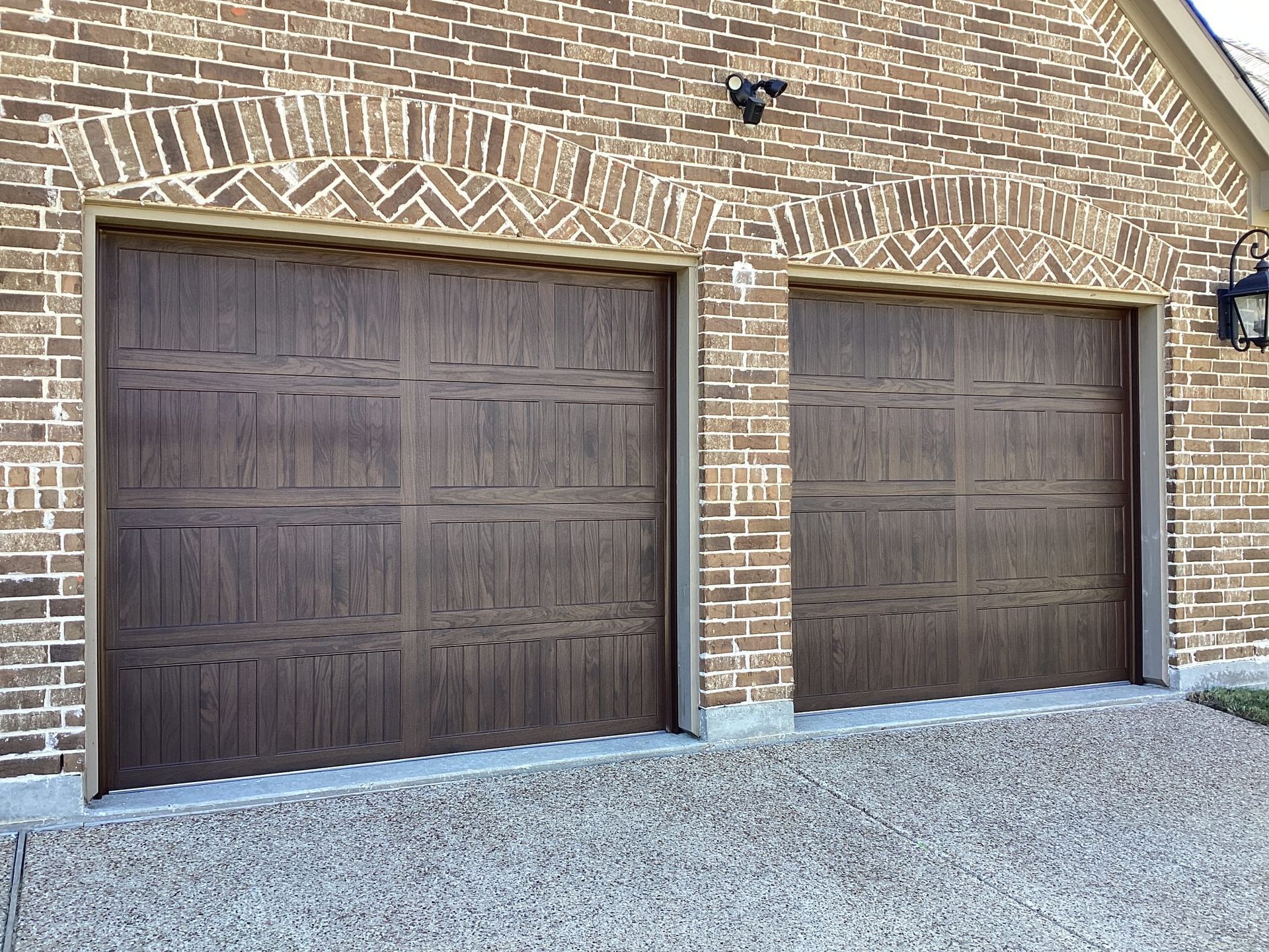 Two dark wood-textured, paneled garage doors set in a tan brick wall with arched brickwork above each door.