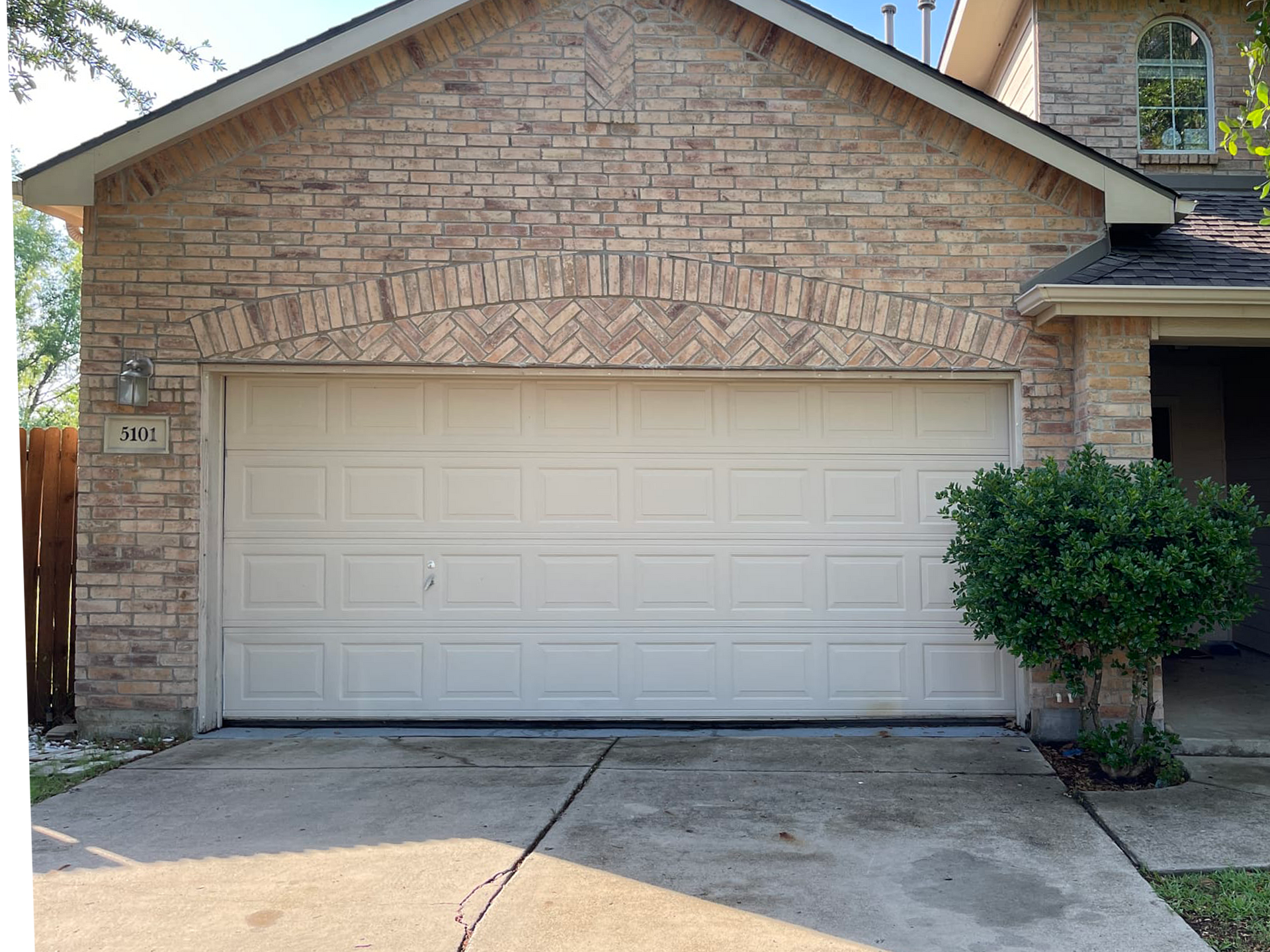Beige garage door on a brick house with an arched brick design above the door. A small bush is to the right.