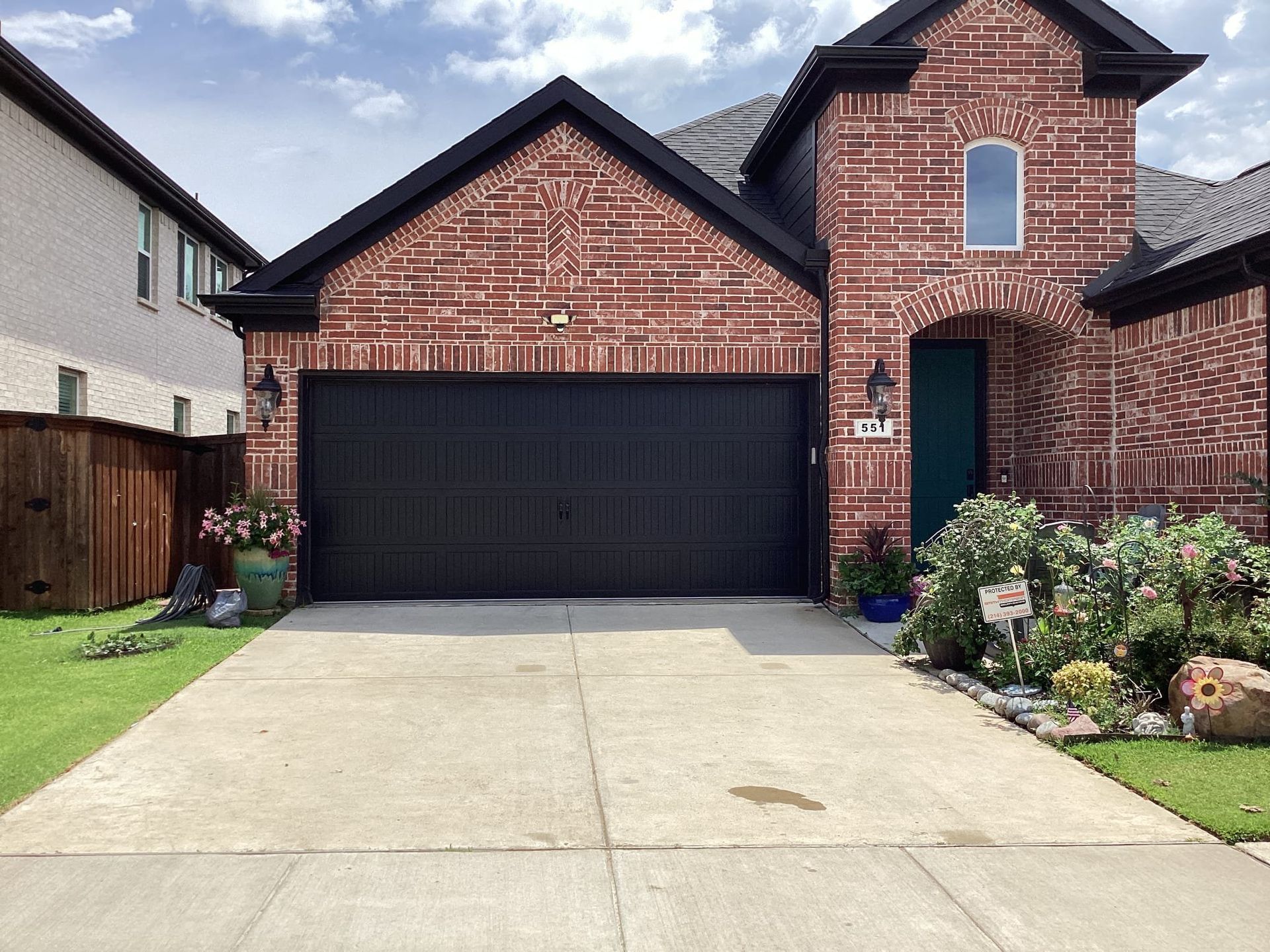 A red brick house with a large black garage door and a concrete driveway, under a blue sky with scattered clouds.