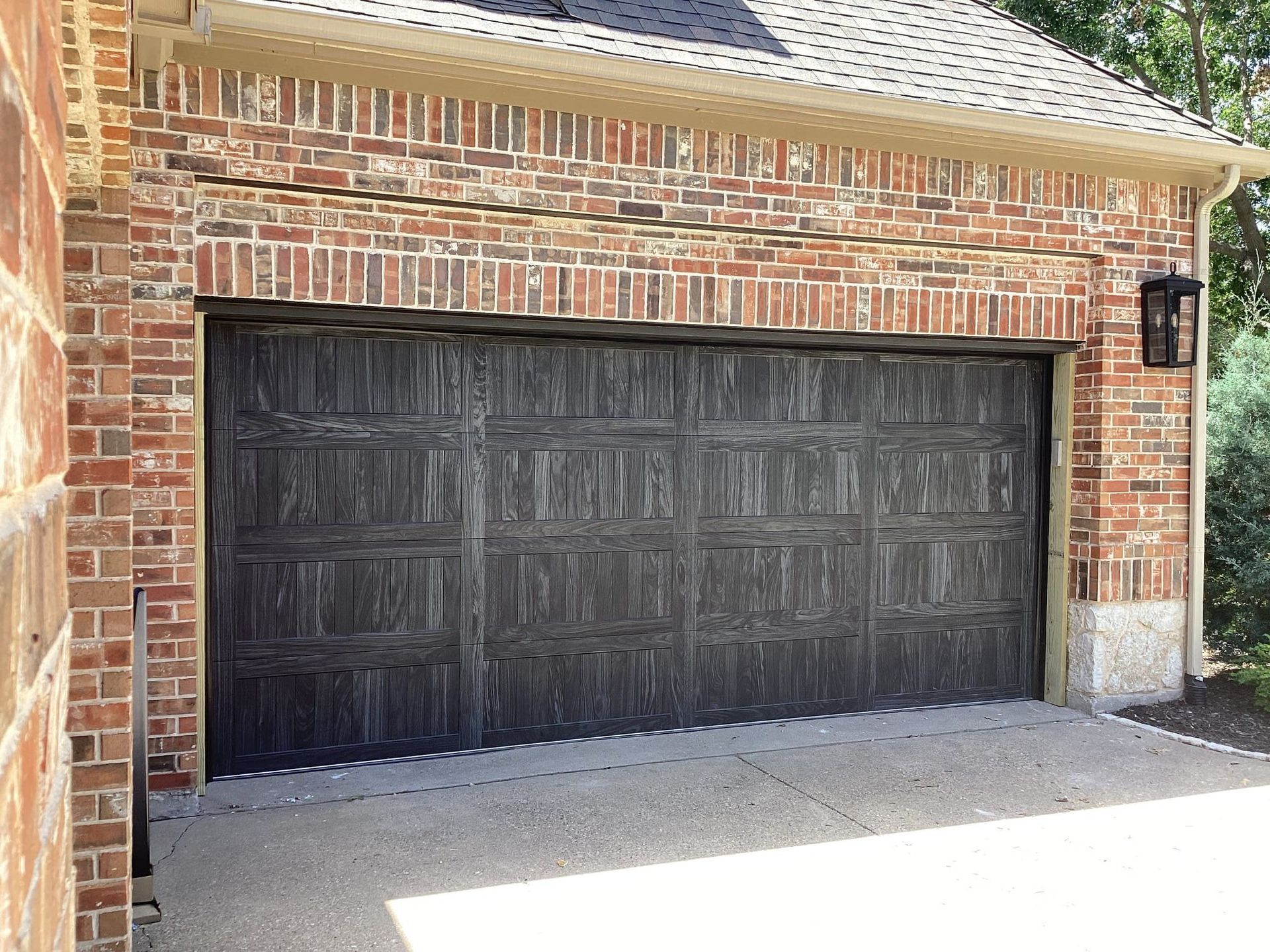 Dark brown, paneled garage door set in a red brick exterior with an arched brick lintel and a black wall lantern.