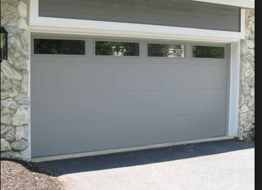 A modern, light gray garage door with four rectangular windows across the top, set between stone-textured exterior walls.
