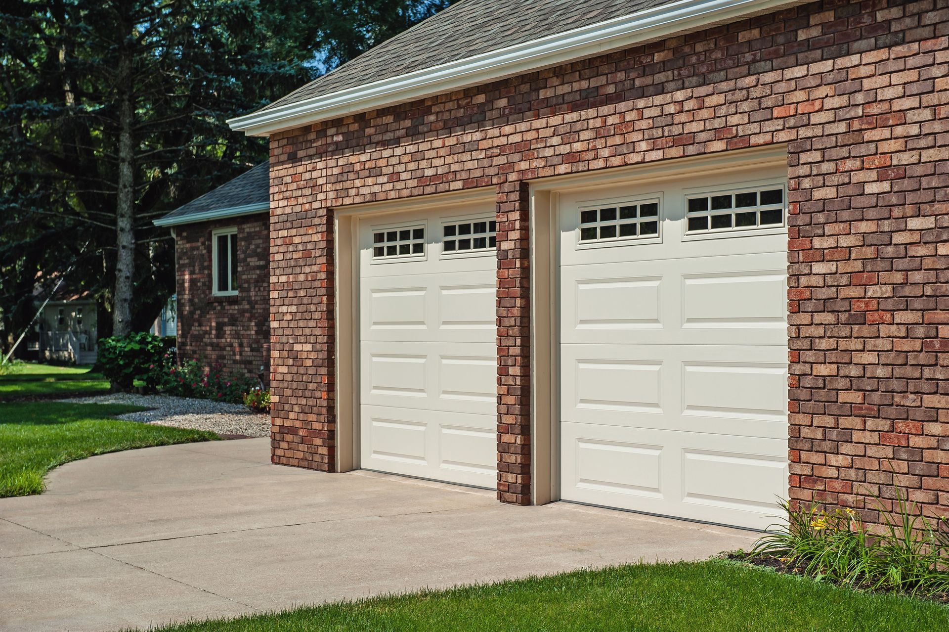 Two beige garage doors with windows, set in a brick residential building next to a concrete driveway.