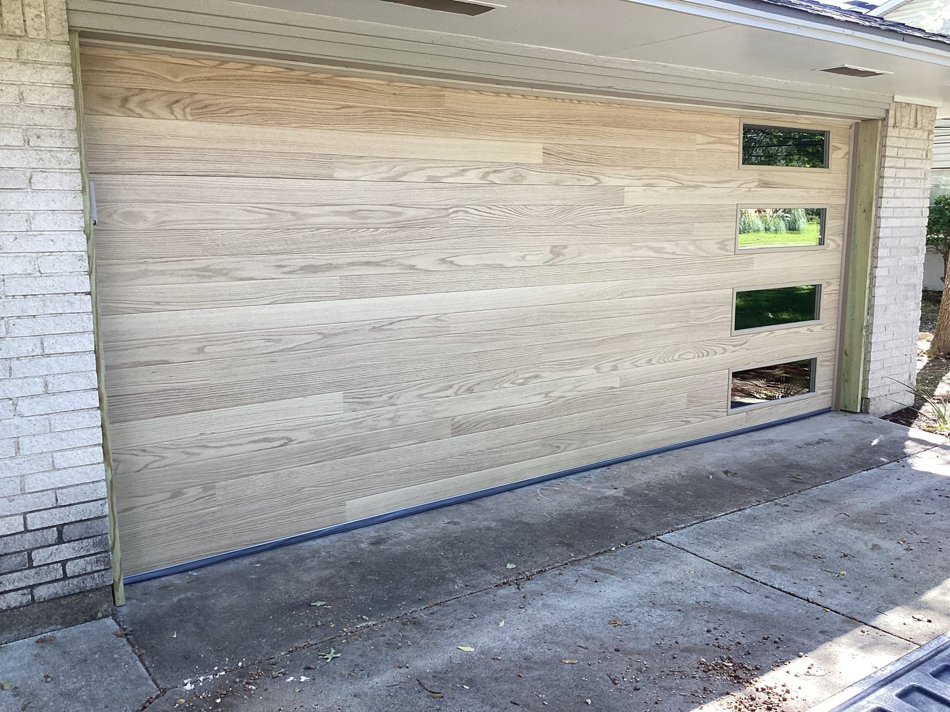 A light-colored wooden garage door with a vertical stack of four rectangular glass windows on the right side.