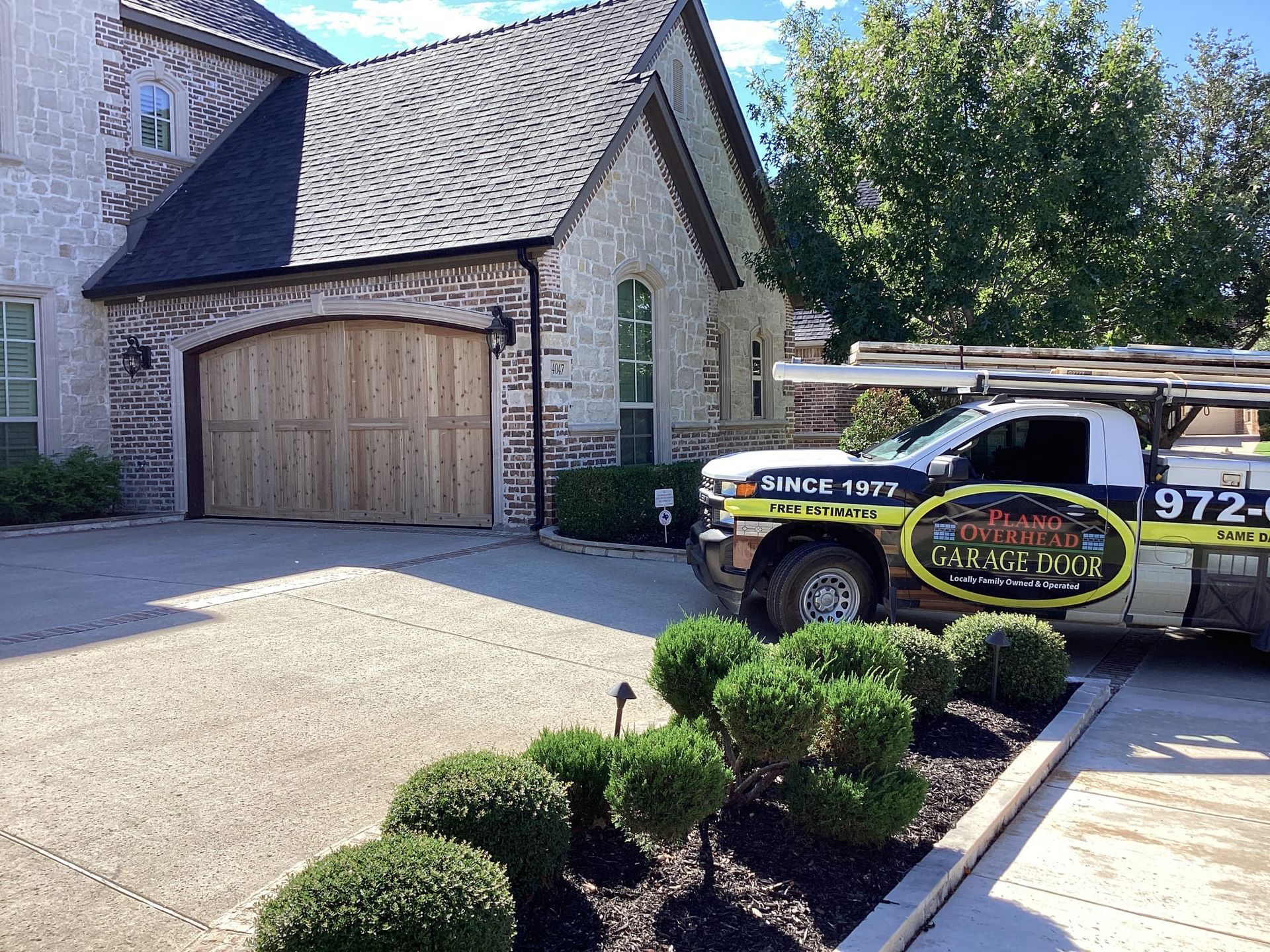A professional garage door service truck parked in the driveway of a brick home with a wooden arched garage door.