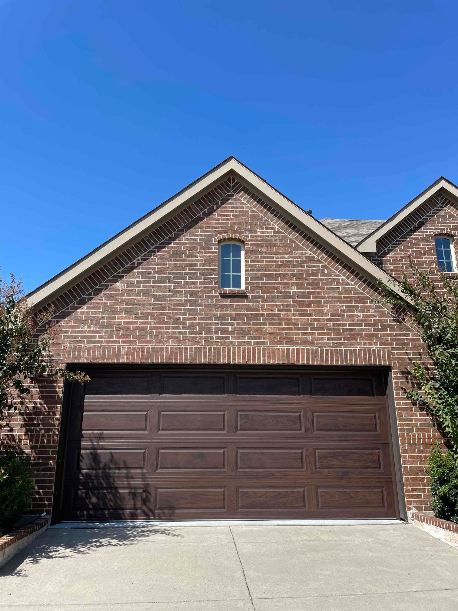 A brown brick house features a dark, paneled garage door beneath a triangular gable with a single, centered window.