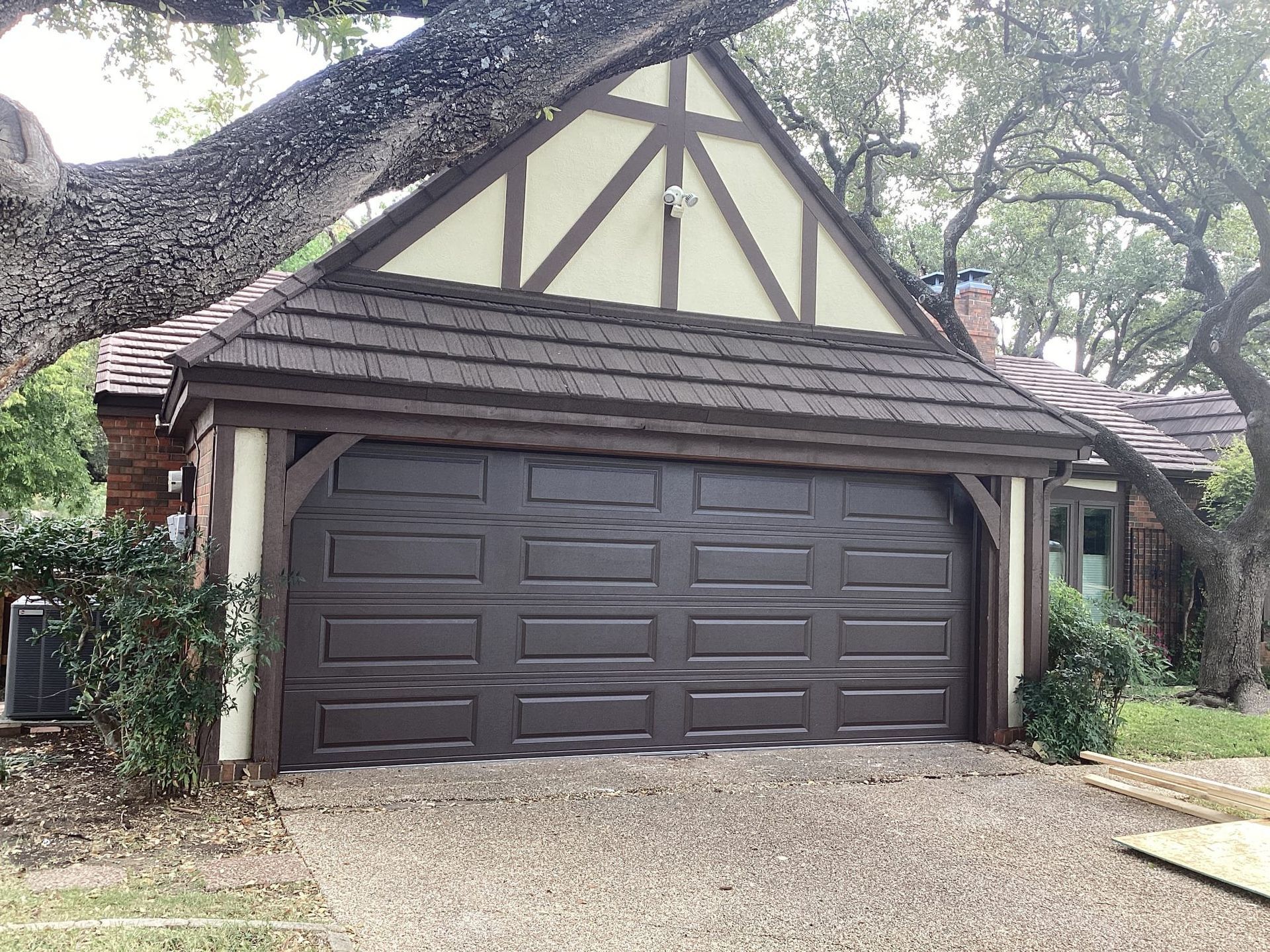A detached garage with a dark brown door, Tudor-style trim, and a gravel driveway, framed by a large tree.