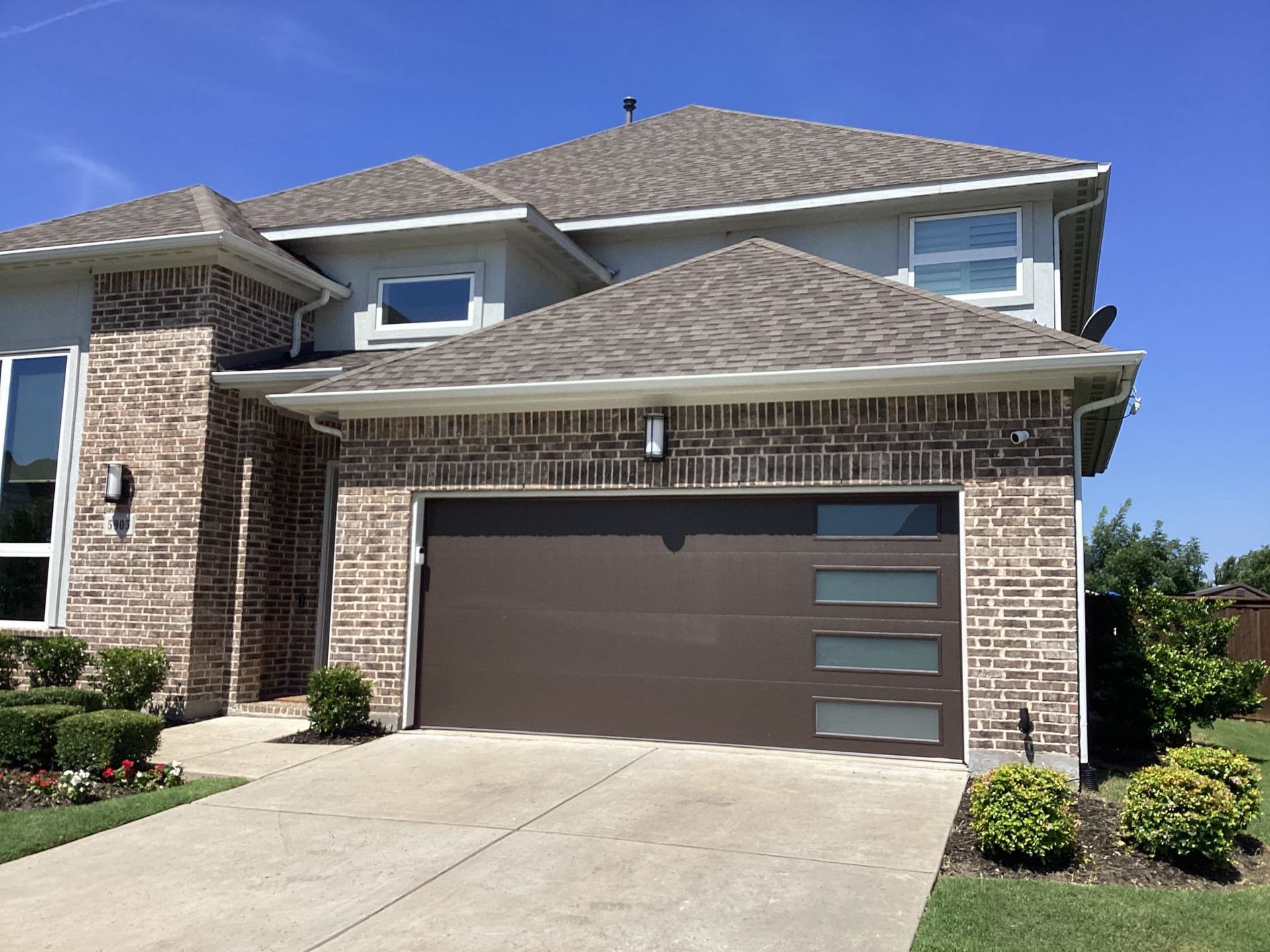 A two-story suburban house with brick and gray siding, a brown modern garage door with glass panels, and a blue sky.