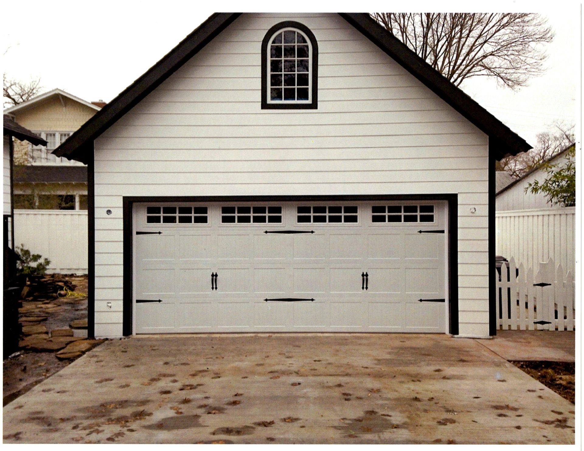 A white detached garage with a black-trimmed arched window and a horizontal panel garage door with windows on a driveway.
