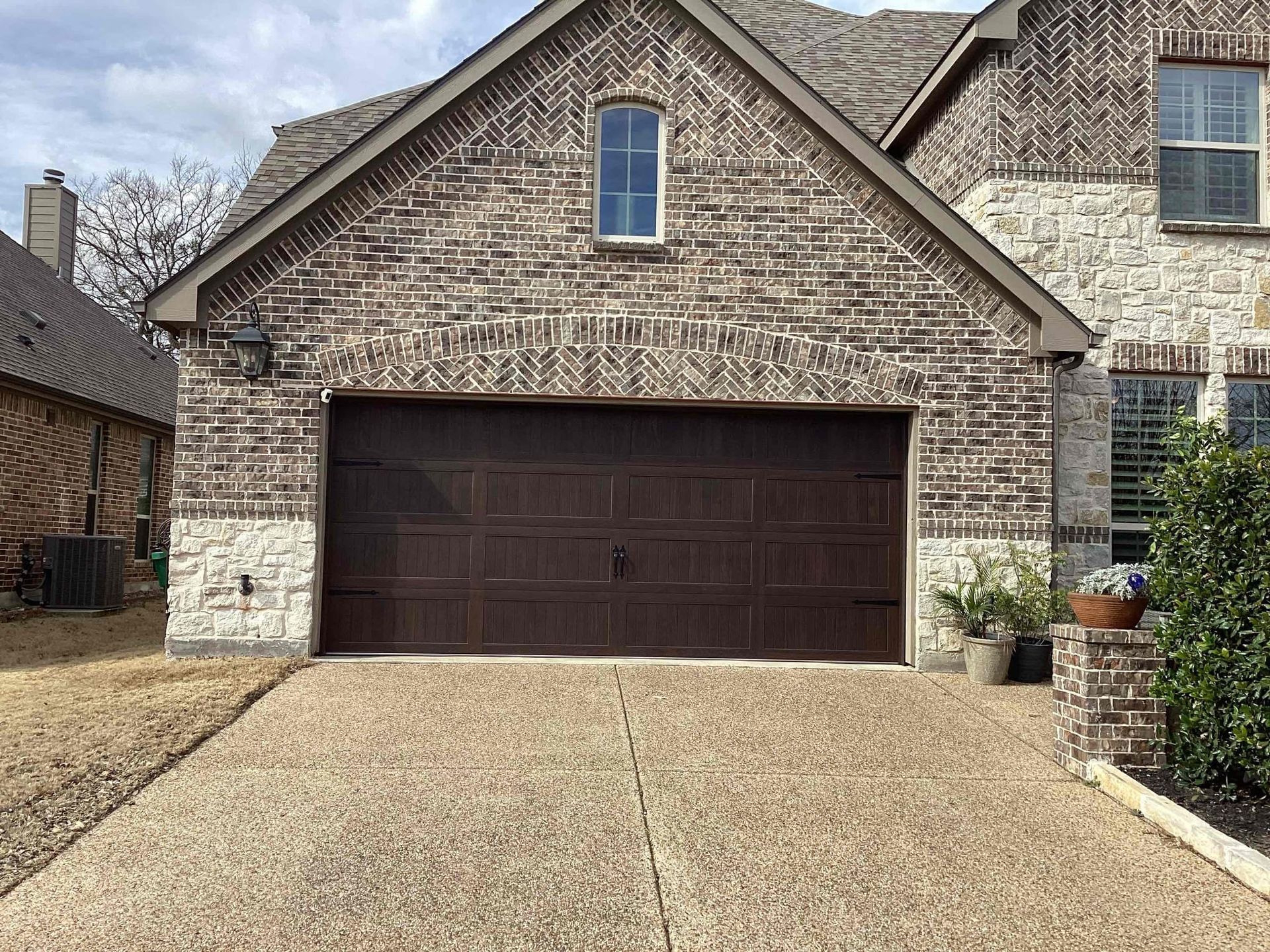 A two-car garage with a brown door, stone accents, and a brick facade under a peaked roof, with a paved driveway.