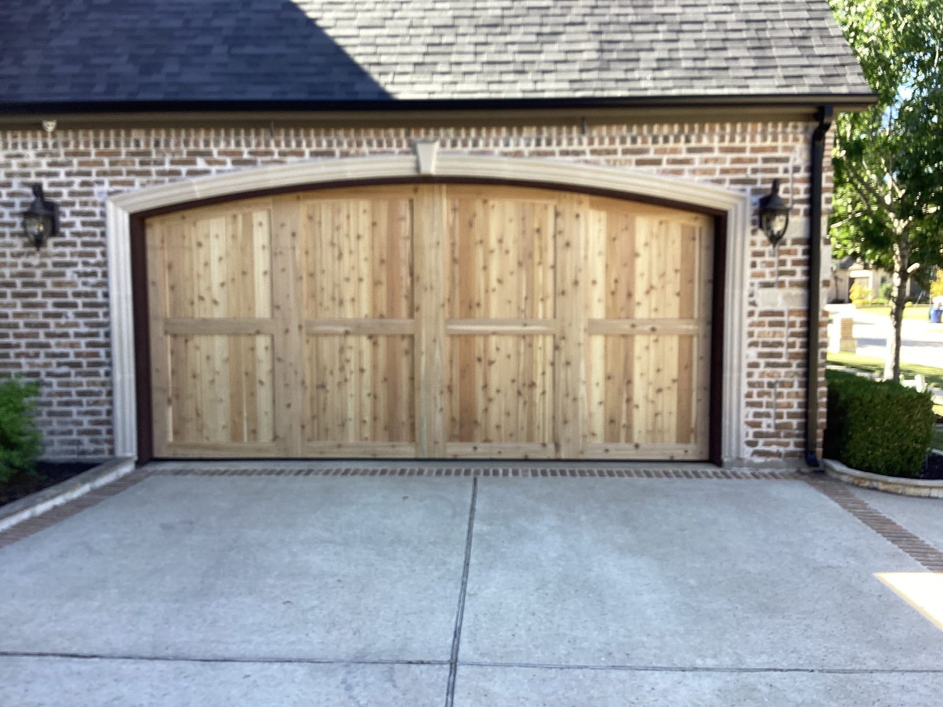 A single-car garage with a light-colored wooden door set in an arched brick frame under a dark shingled roof.
