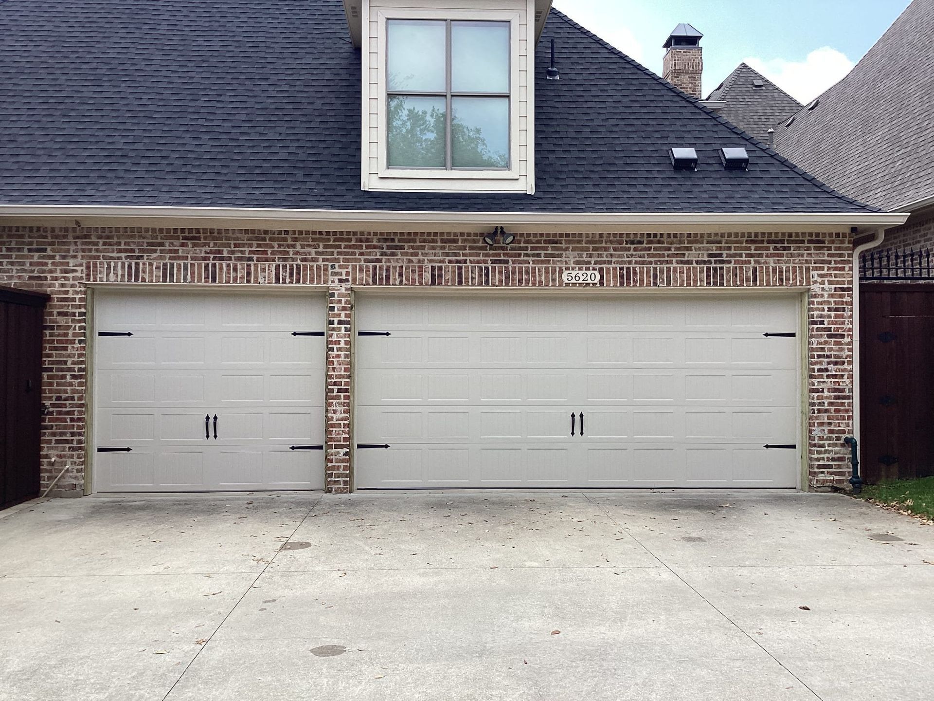 A two-car garage with cream-colored paneled doors and decorative black hinges set in a brick exterior with a roof dormer.