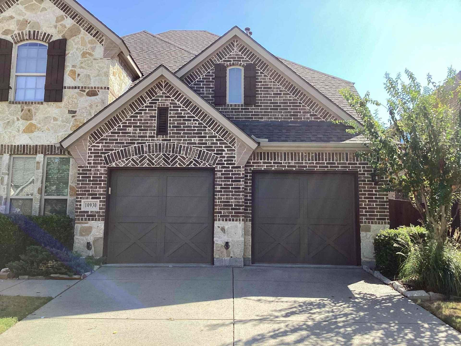 A two-car garage with brown doors on a brick and stone suburban house under a clear blue sky.
