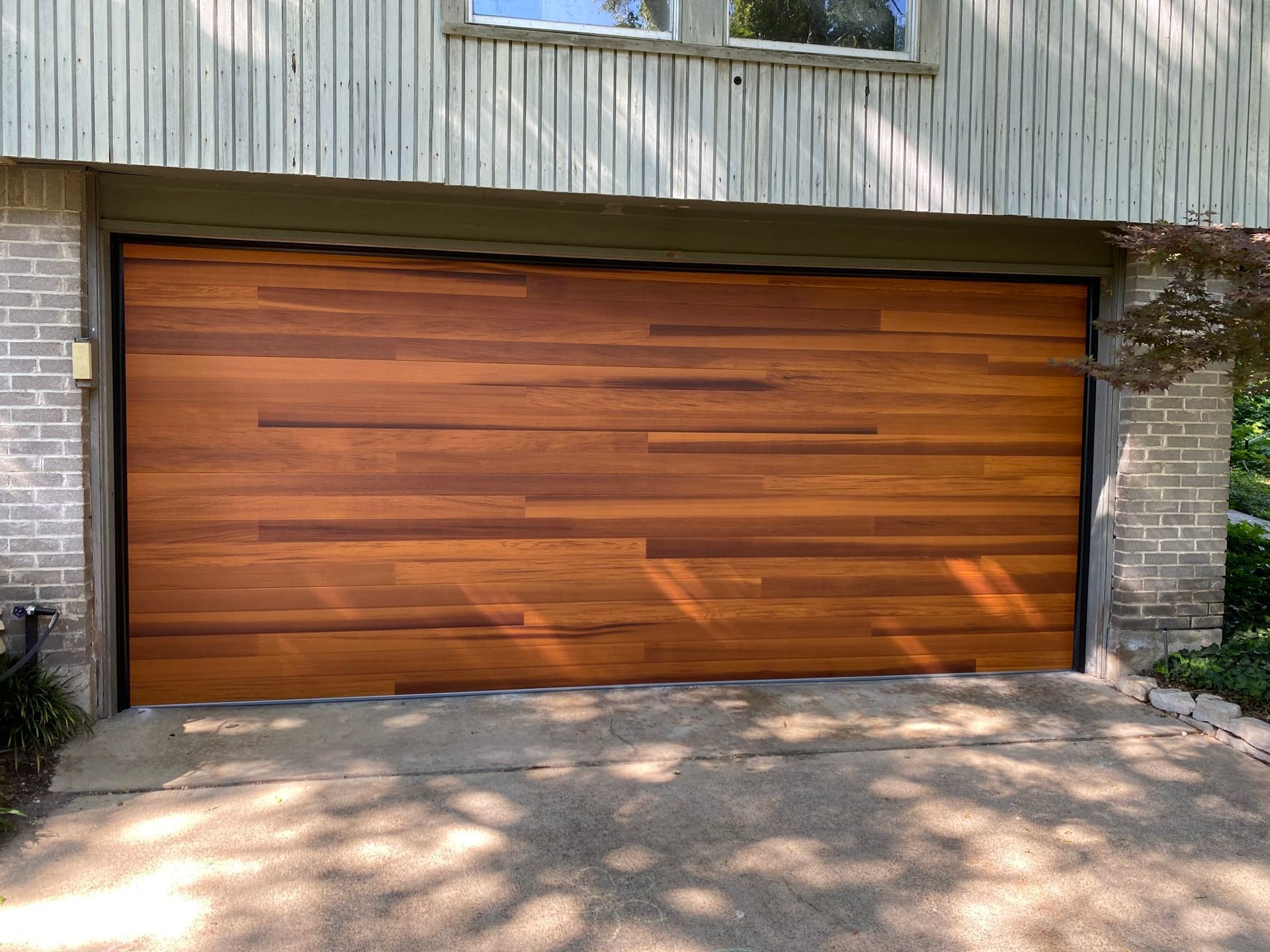 A modern, wide garage door with horizontal, natural wood planking, set in a light brick and vertical siding exterior.