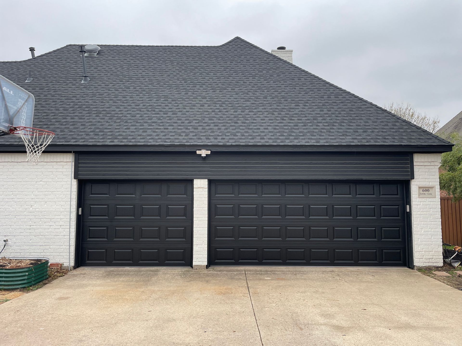 A house exterior featuring a dark grey shingled roof, white brick walls, and two matching black paneled garage doors.
