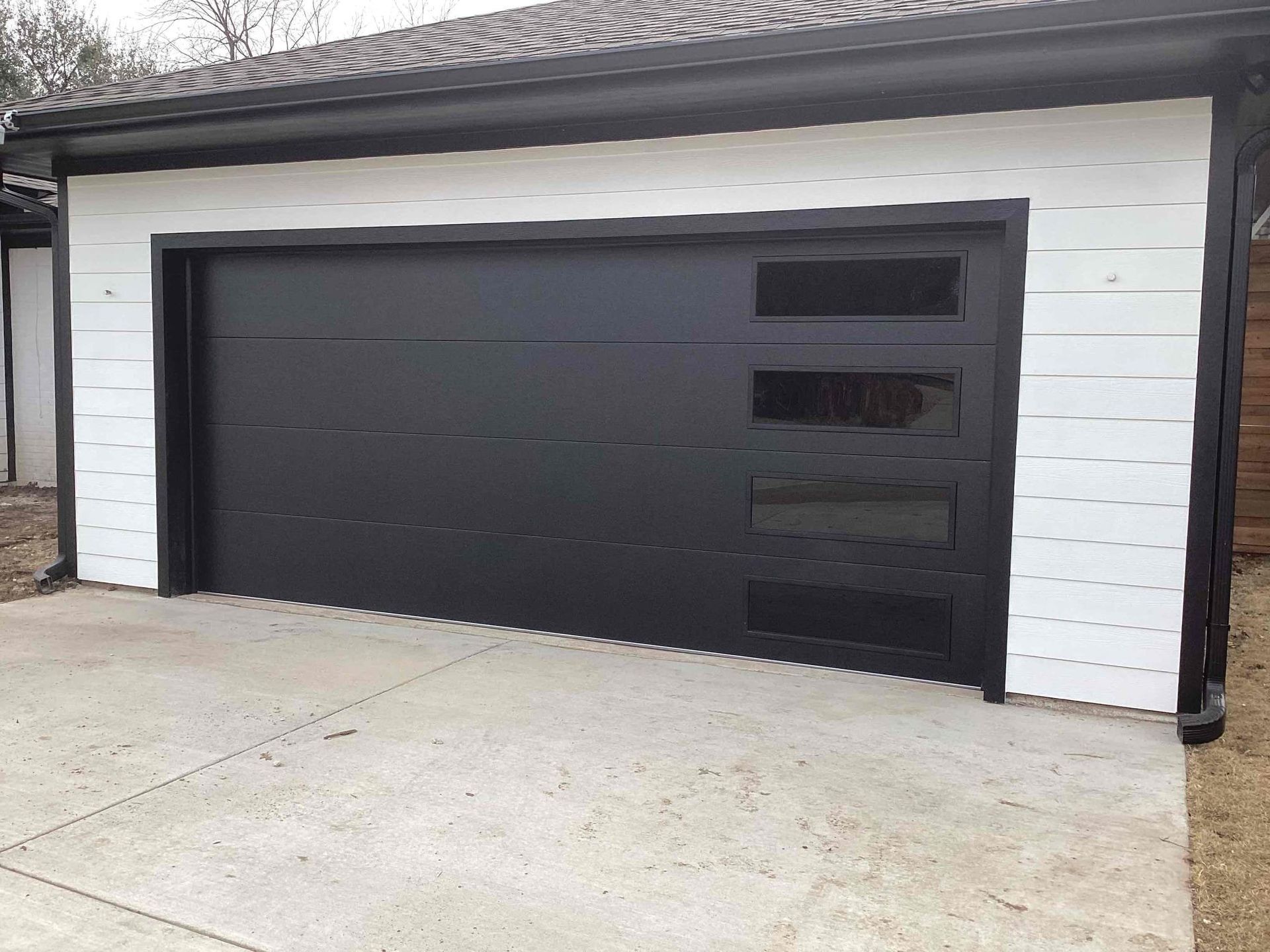 Modern black garage door with side windows installed in a building with white horizontal wood-style siding.