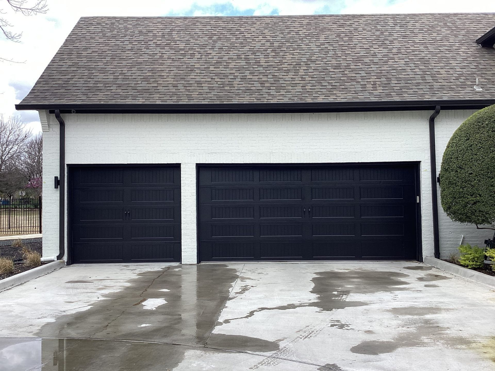 A white brick garage with two black carriage-style doors and a dark shingled roof, seen from a wet concrete driveway.