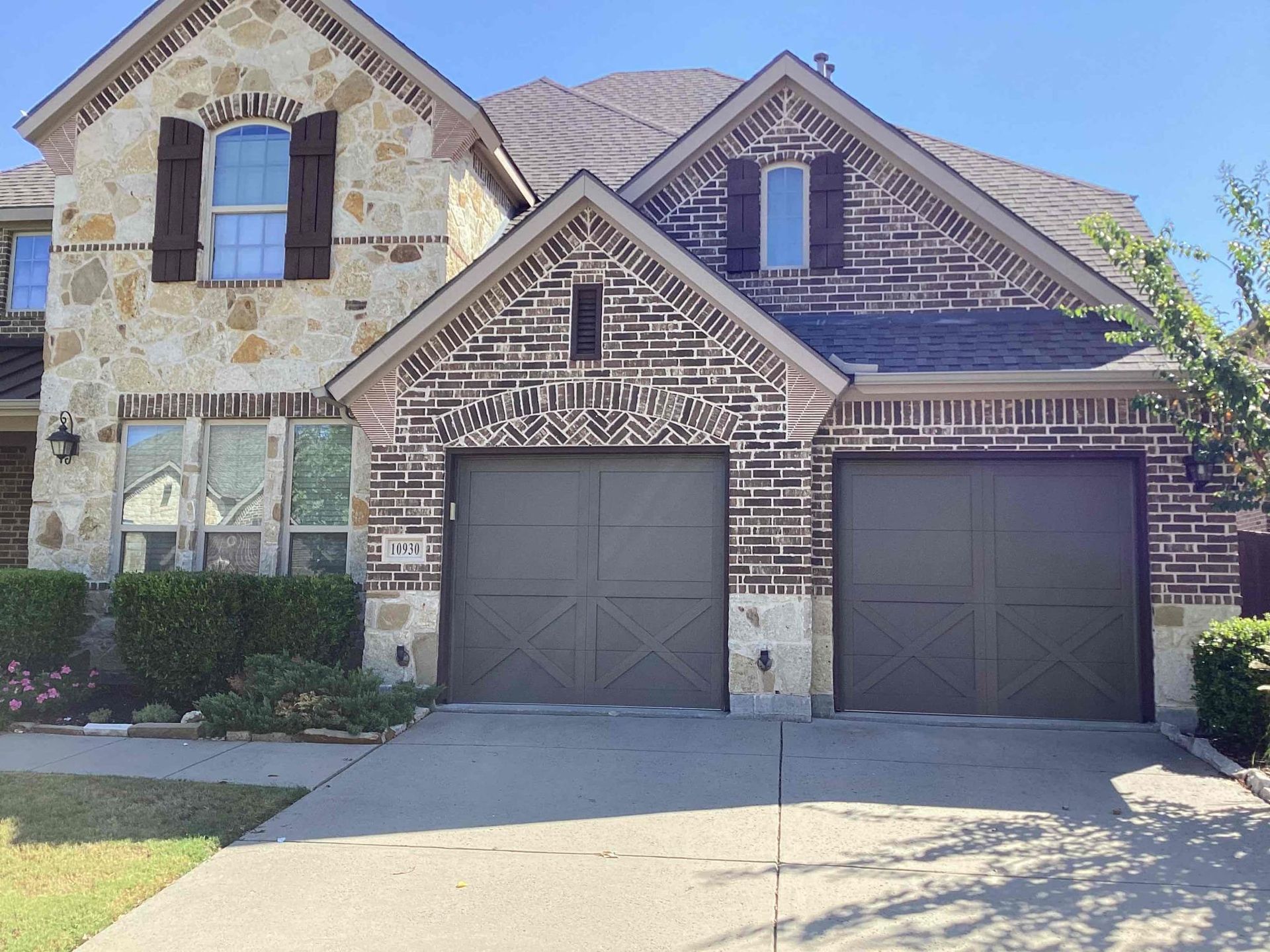 A suburban house exterior with a stone and brick facade, two brown garage doors, and a small front lawn on a sunny day.