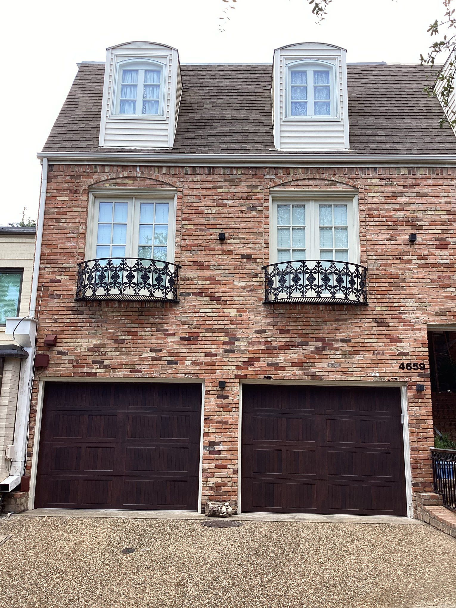 A three-story brick townhouse featuring two dark brown garage doors, two windows with decorative railings, and two dormers.