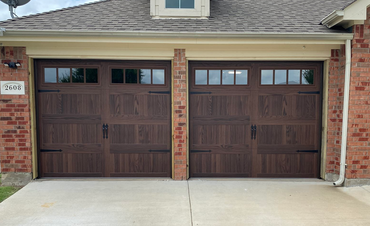 Two brown wooden garage doors with windows, in a brick house, on a concrete driveway.