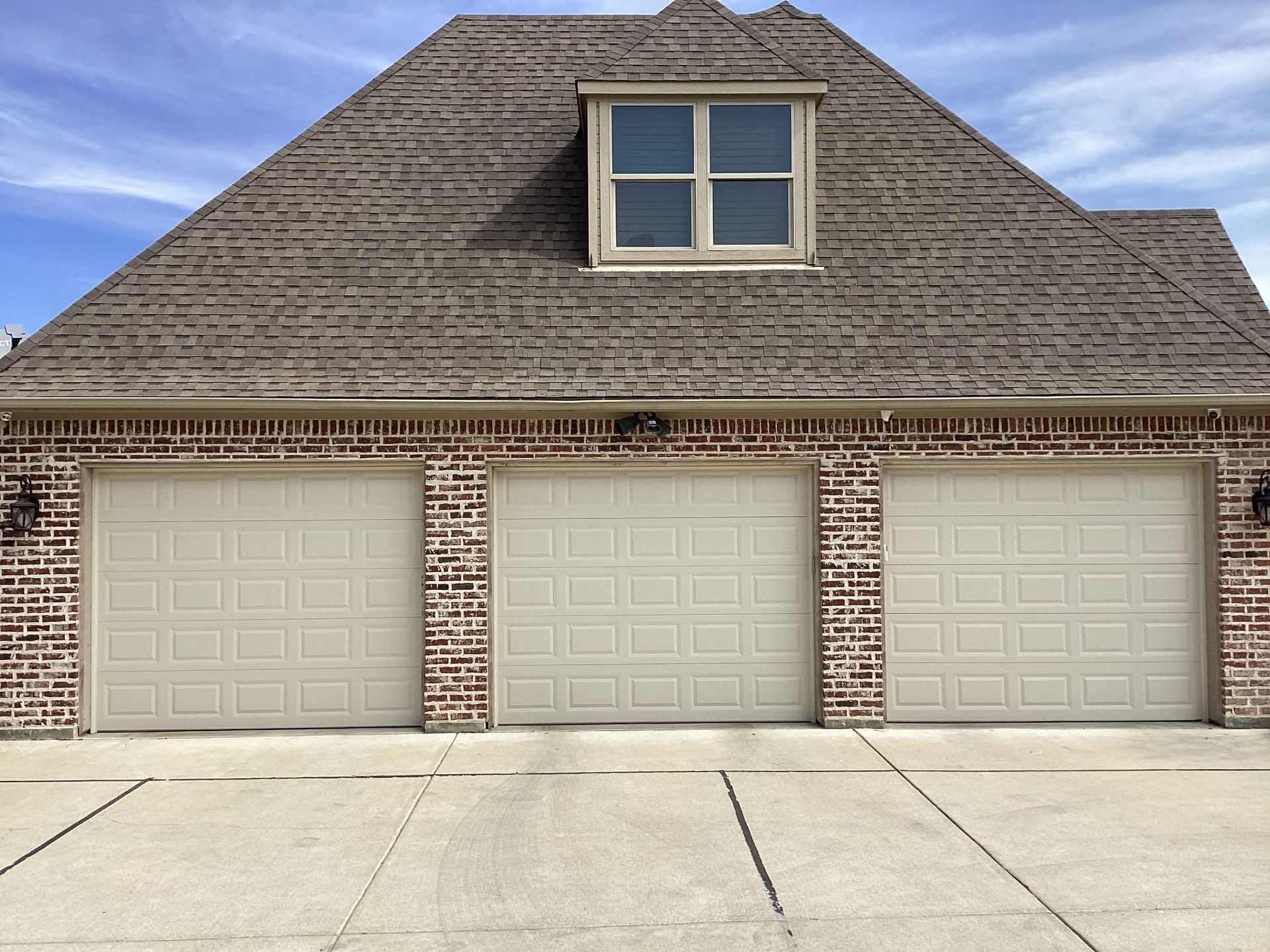 Three beige garage doors set into a brick exterior wall with a dormer window above on a shingled roof.