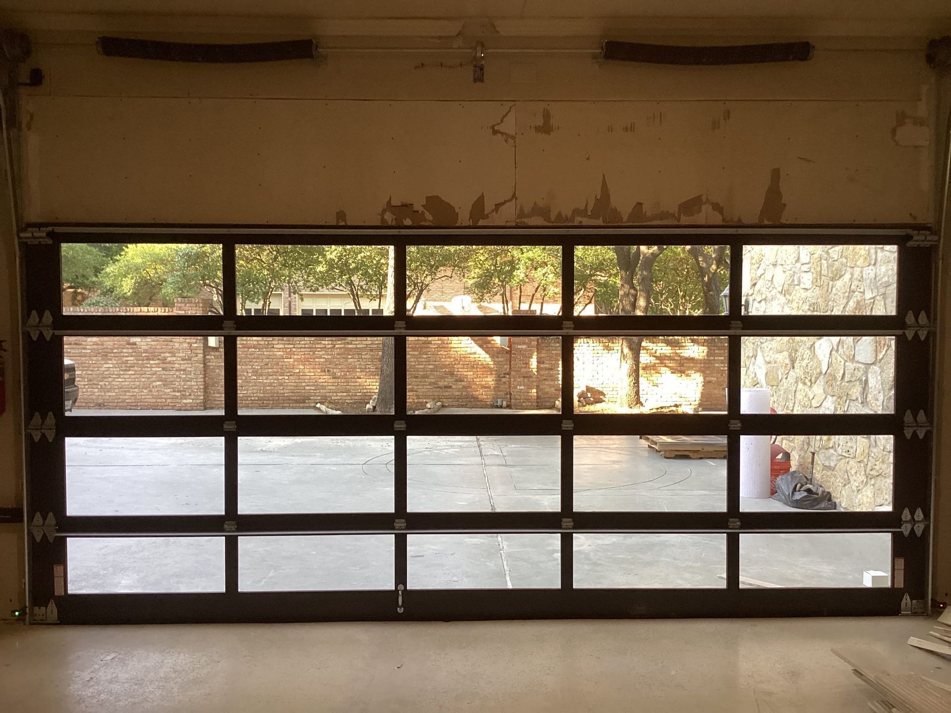 A modern, black-framed glass garage door with four horizontal panels, looking out onto a paved driveway and trees.