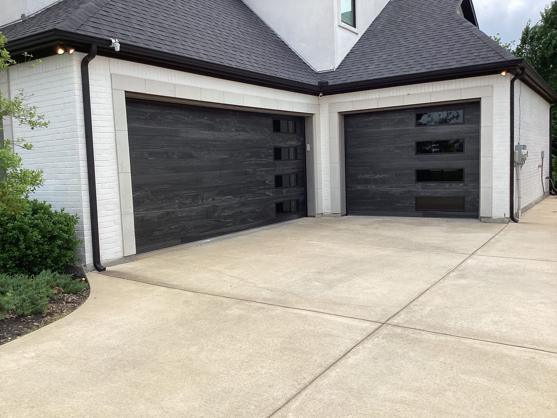 Two dark modern garage doors with horizontal windows installed in a white brick home with a concrete driveway.