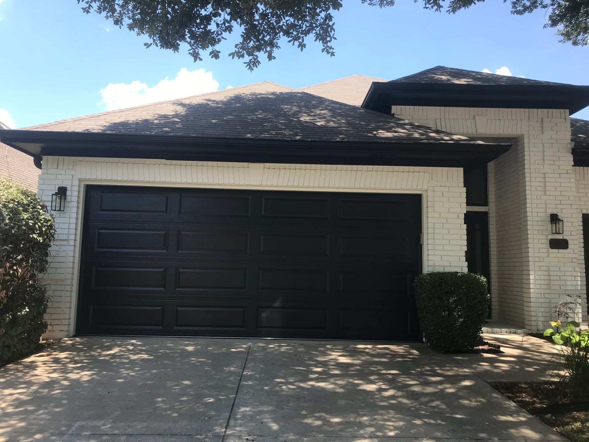 A white brick house with a large black garage door and a dark shingled roof under a bright, sunny sky.