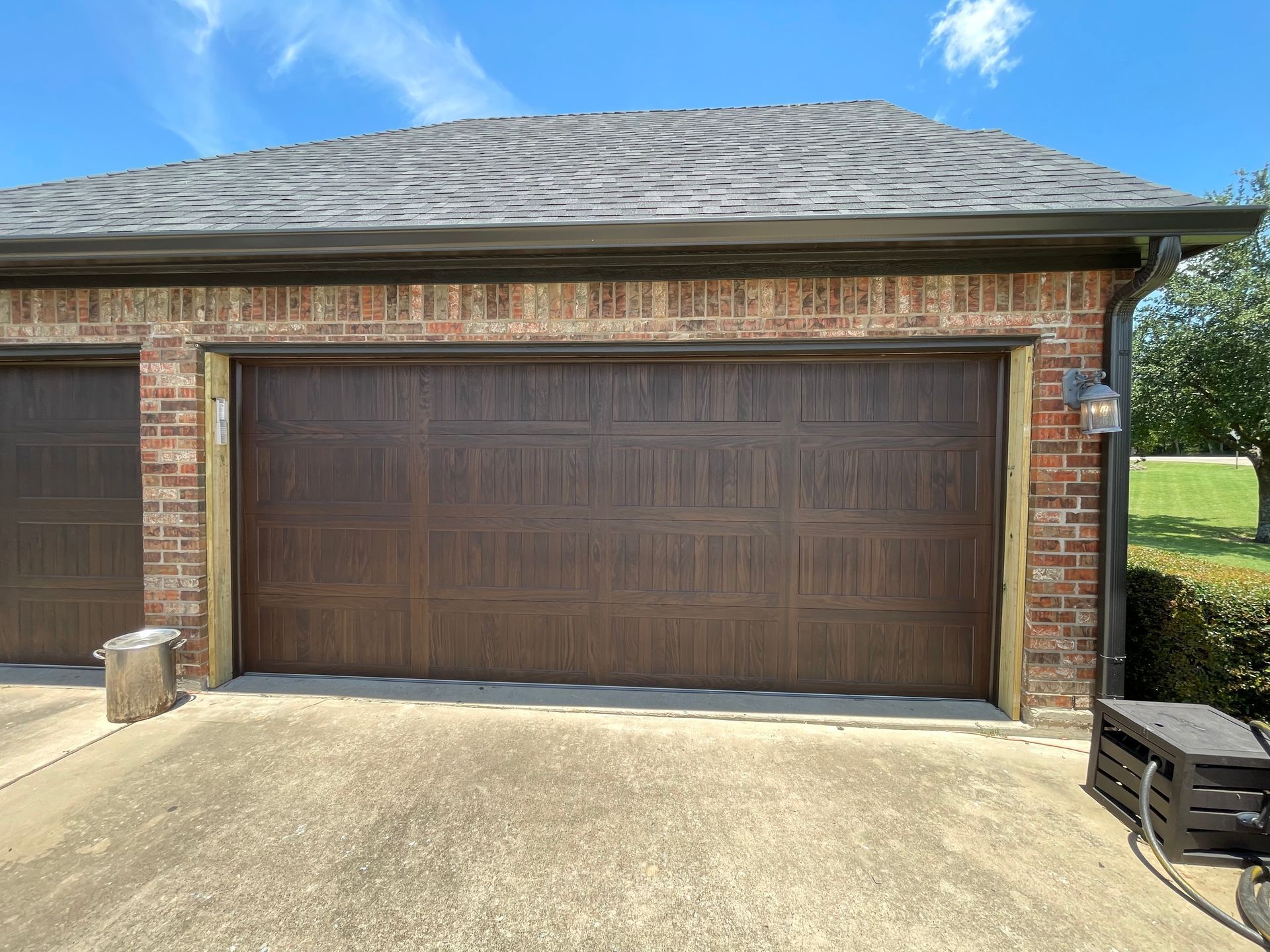 A brick garage exterior with a dark brown, multi-panel garage door under a shingled roof on a sunny day.