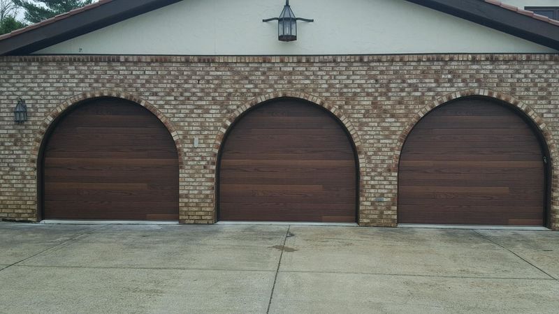 A three-car garage with arched, dark wood doors set into a beige brick exterior wall with two exterior wall lamps.